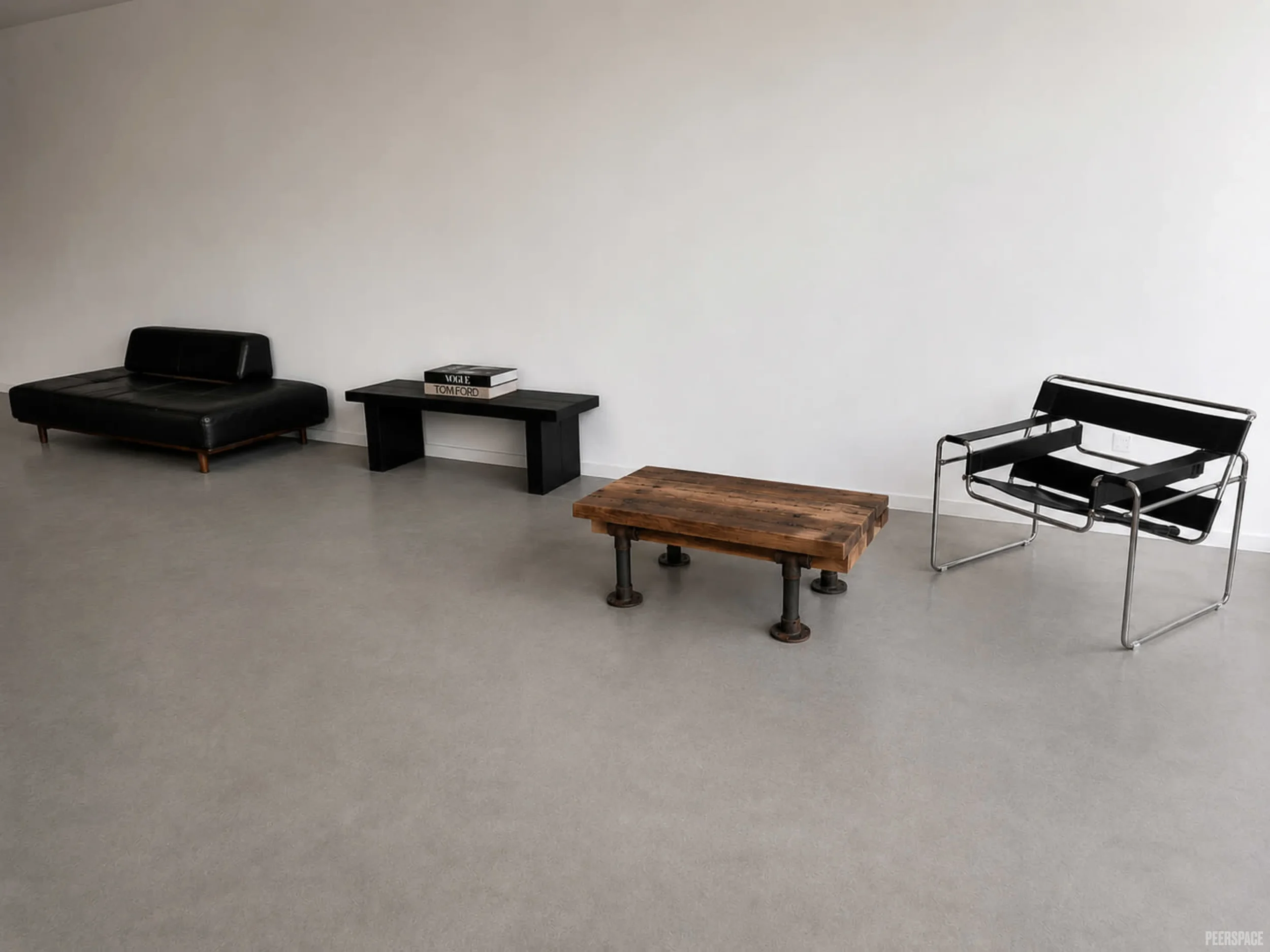A minimalist room with a black leather bench on wooden legs, a black coffee table, a small black side table with two books, and a black and chrome chair, all against a plain white wall.