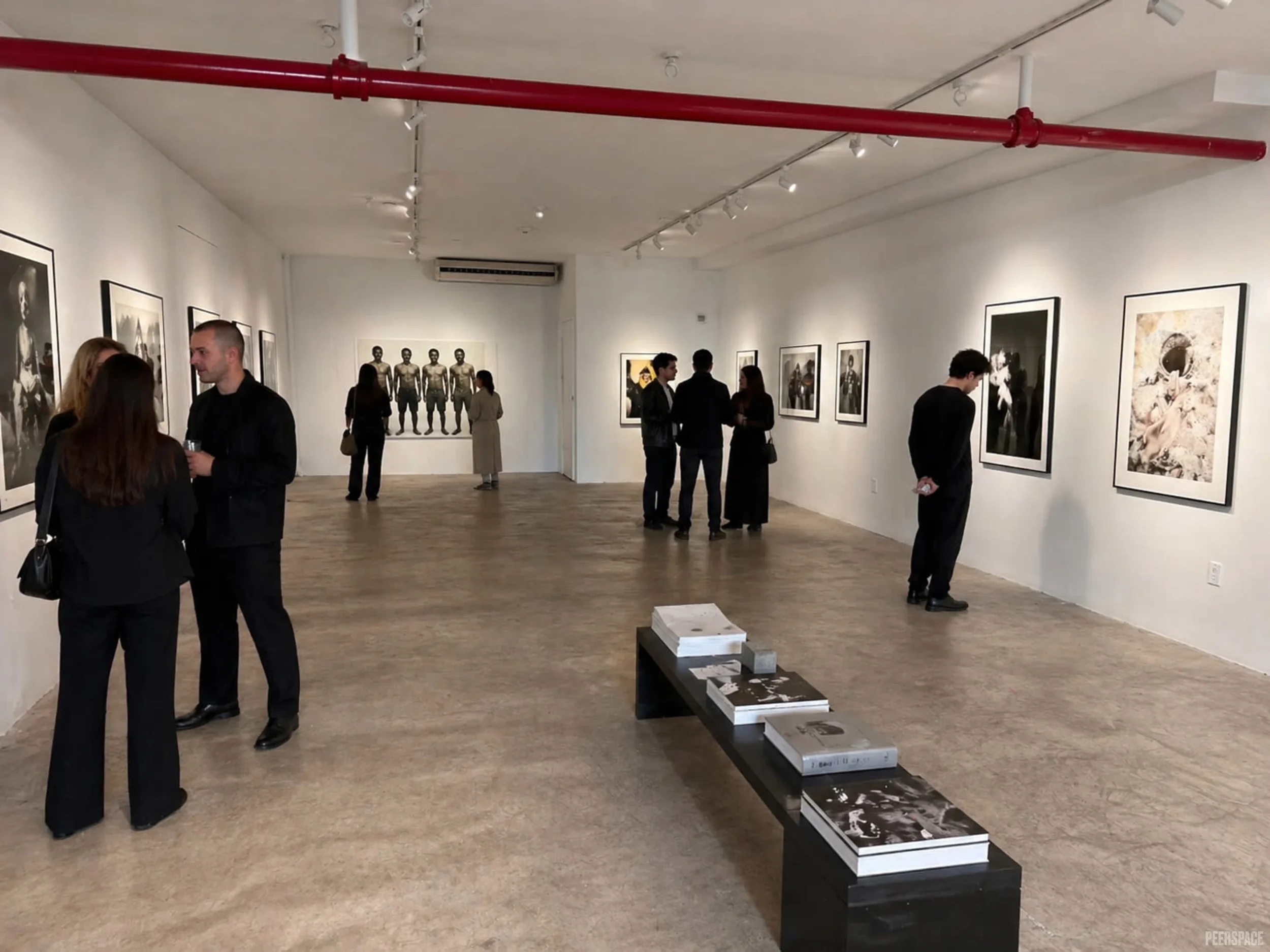 People viewing black and white artwork in an art gallery with photographs on the wall, red pipe running along the ceiling, and a wooden floor.