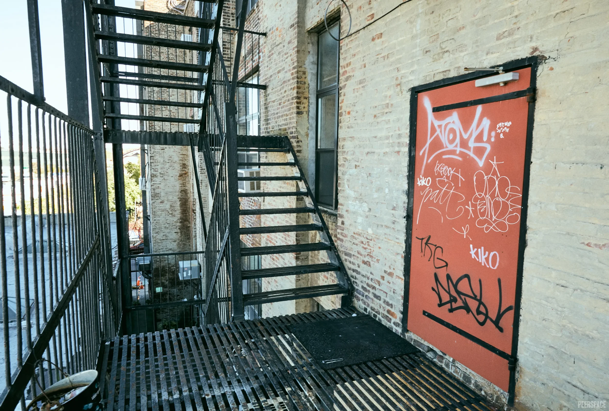 View of a fire escape on a brick building with graffiti-covered red door on the side and black metal stairs and railing.