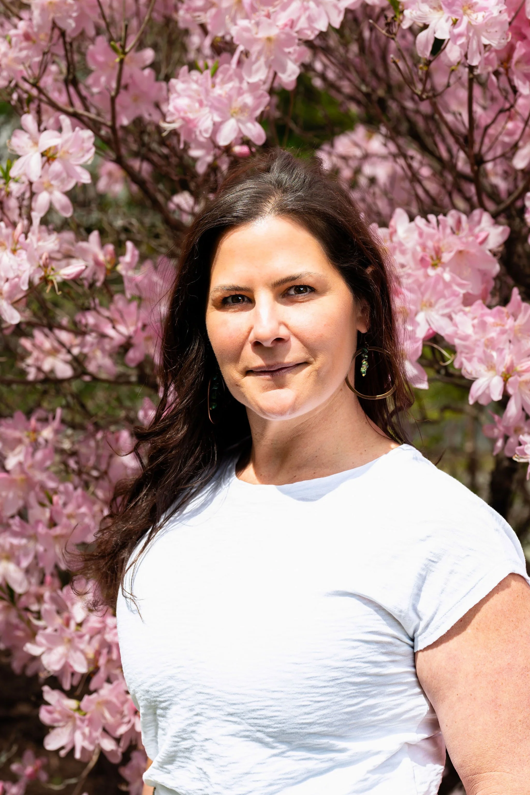 Photo of brunette woman wearing a white shirt standing in front of pink flowering bush