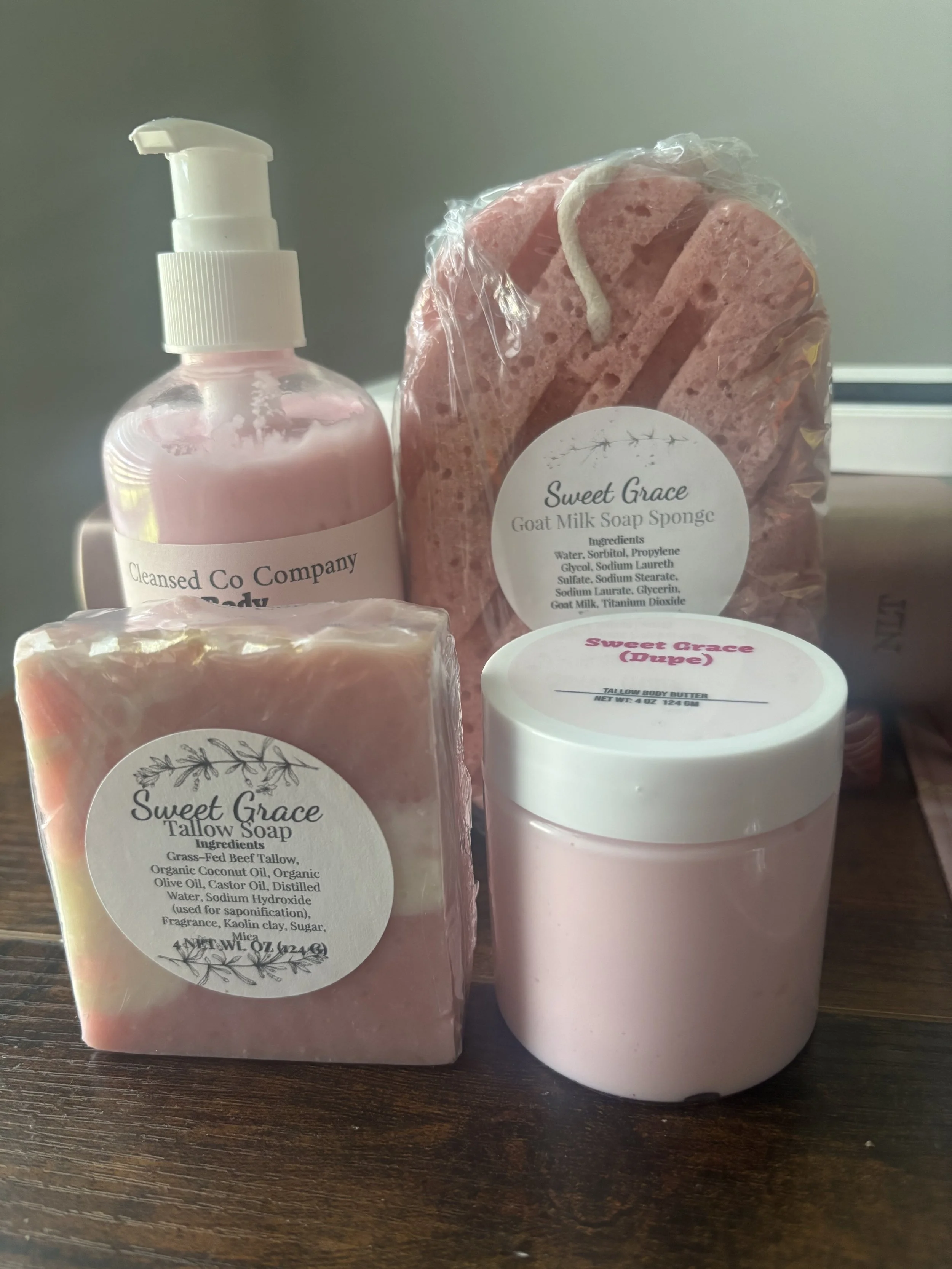 Image shows a pink soap bar, a pink liquid soap bottle with a pump, and a pink body butter container on a dark wooden surface. The soaps are labeled with the brand "Sweet Grace" and ingredients.
