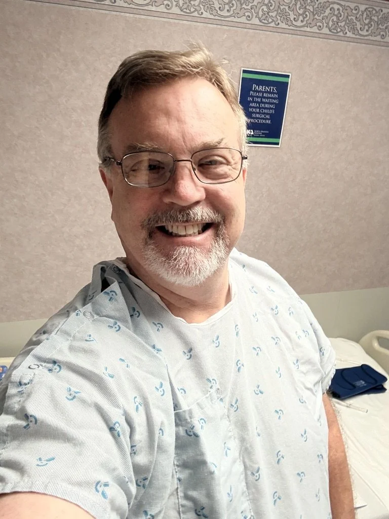 A man in a hospital gown taking a selfie in a hospital room, smiling, with medical equipment and a sign on the wall that reads 'Parents: please remain in the waiting area during your child's surgical procedure.'