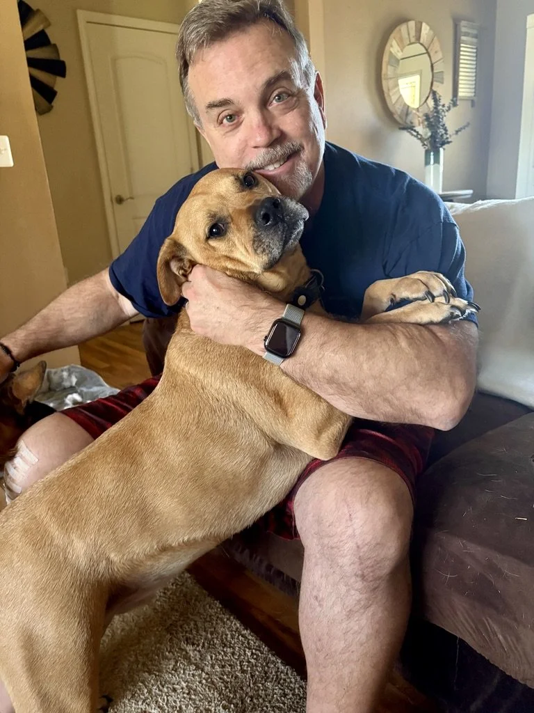A man with a beard and wearing a navy blue shirt hugging a tan dog with a black collar indoors.