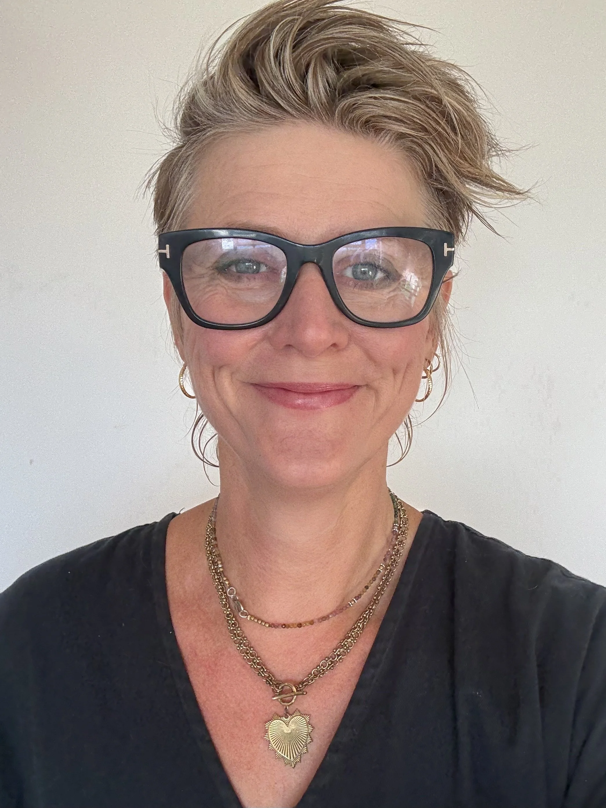 Close-up of a woman with short, wavy blond hair, wearing large black glasses, gold hoop earrings, and multiple gold necklaces, smiling against a plain light-colored background.