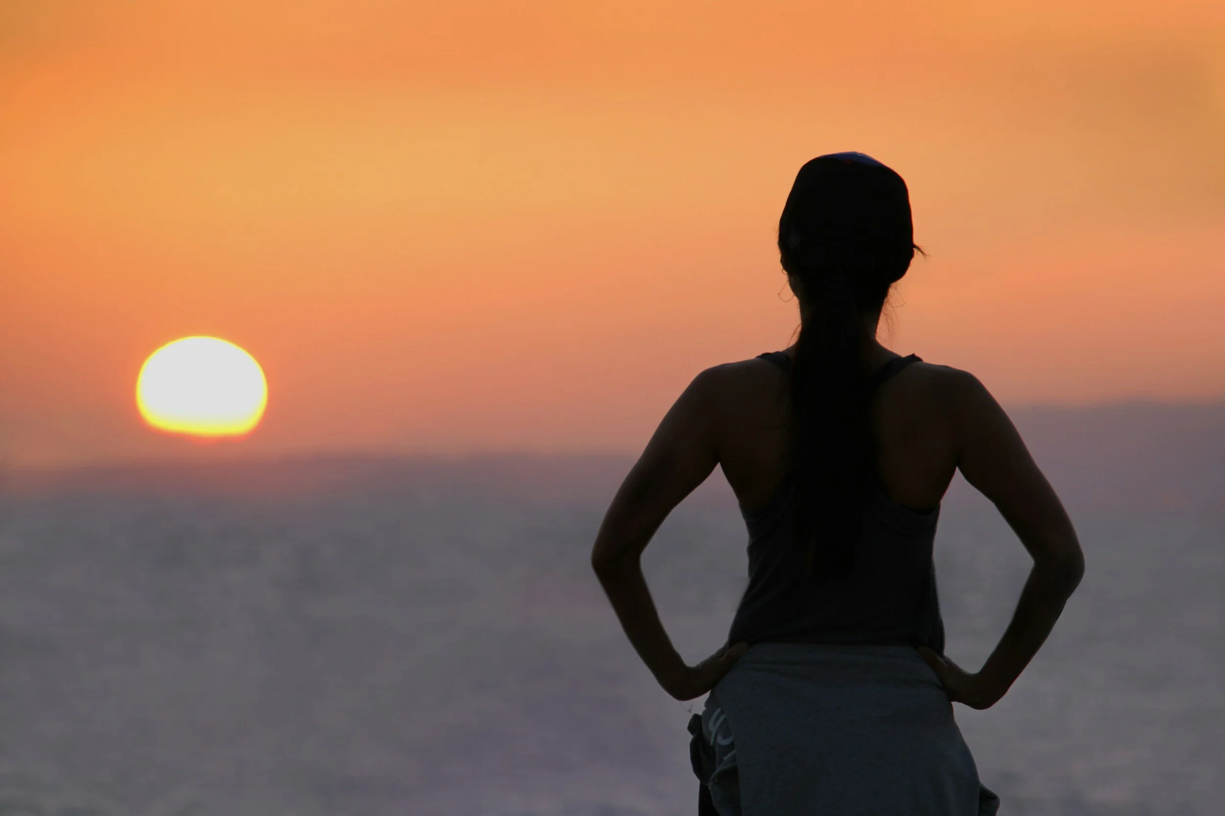 A person practicing yoga or meditation outdoors at sunset, sitting cross-legged with hands on hips, facing the setting sun in the sky.