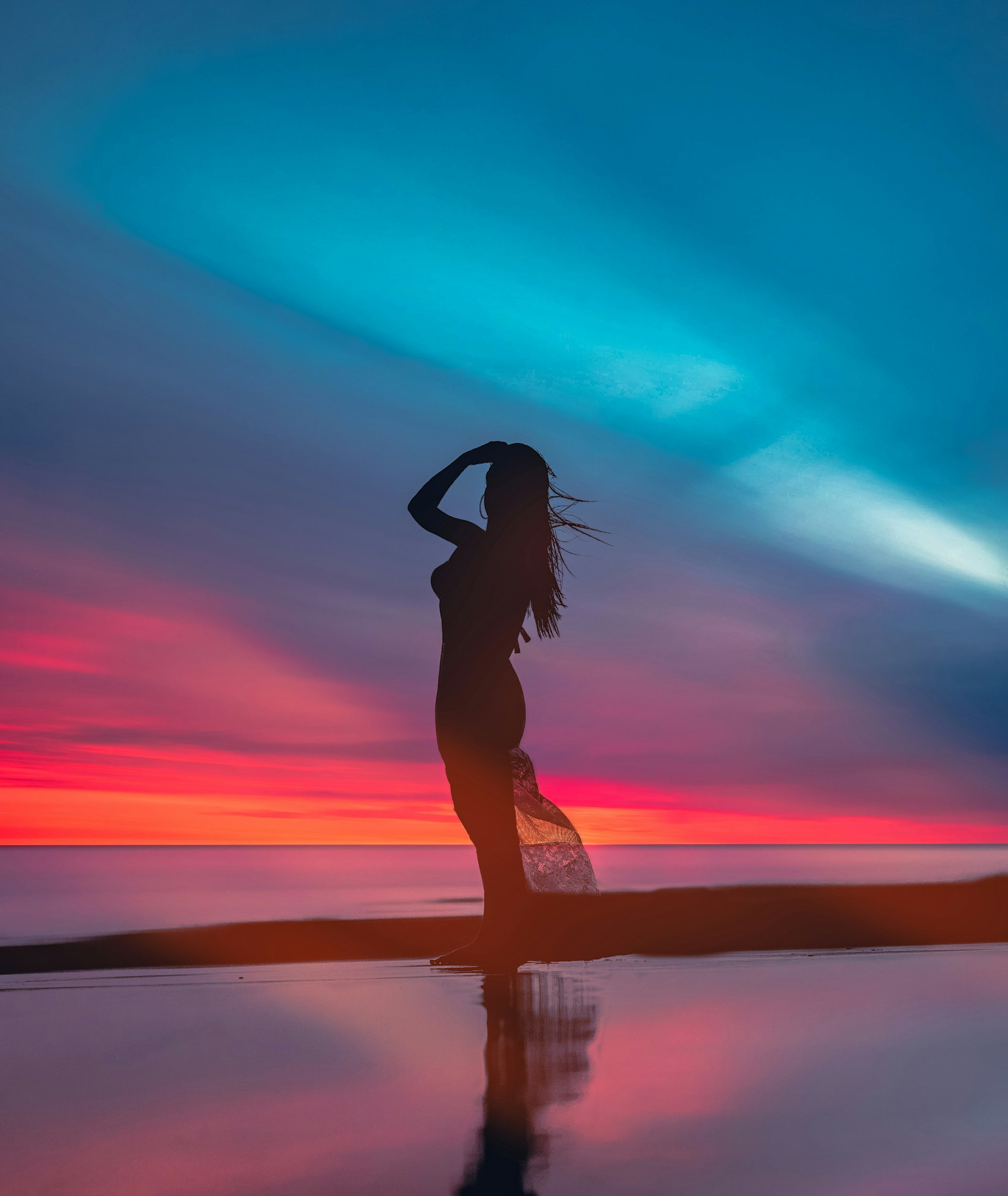 Silhouette of a woman standing on the beach at sunset, with colorful sky and reflection in the water.