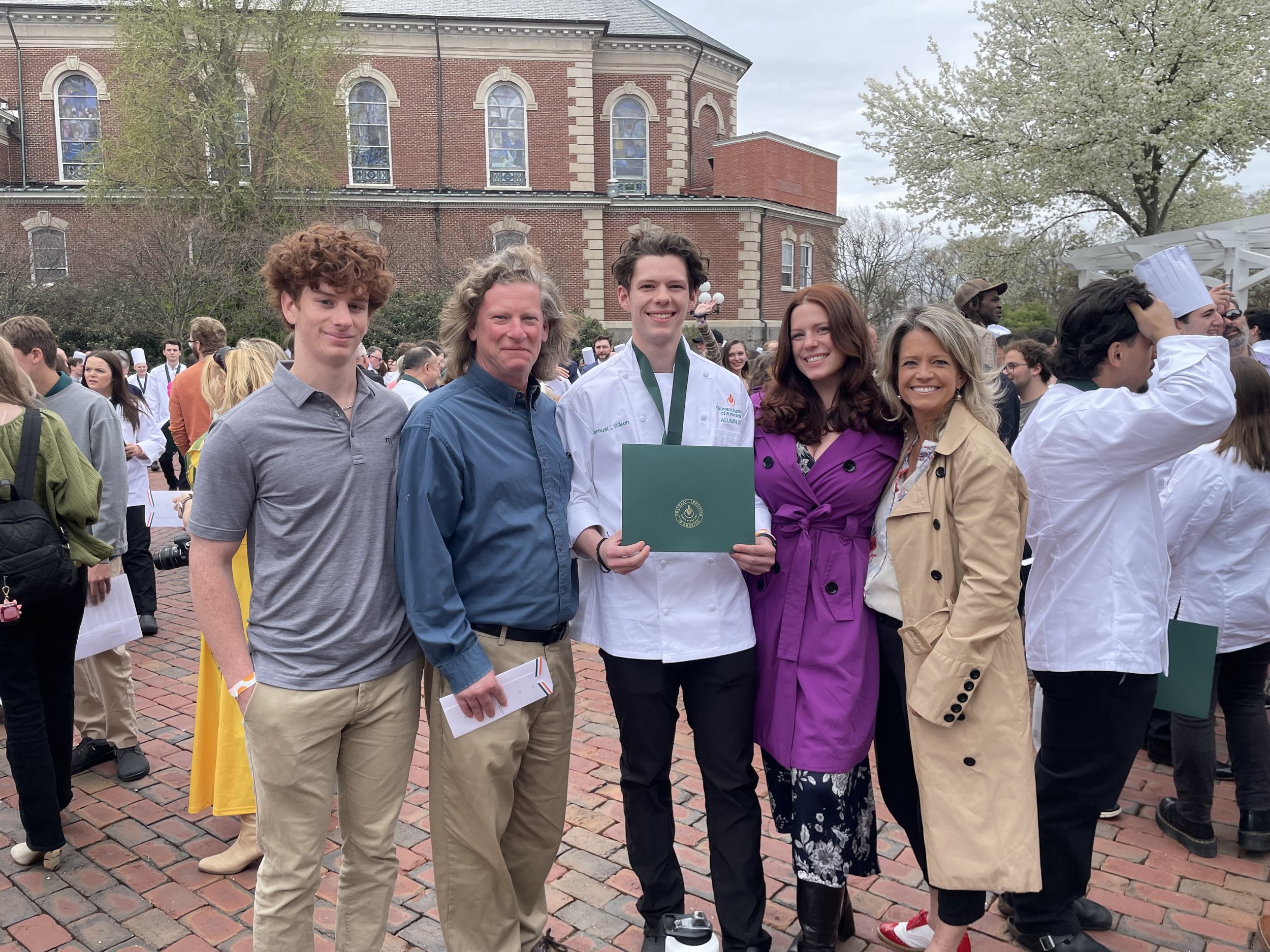 A group of people, including a young man in a chef's coat holding a green certificate with a medal around his neck, stand together outdoors in front of a brick building and trees. The scene appears to be a graduation or award ceremony.