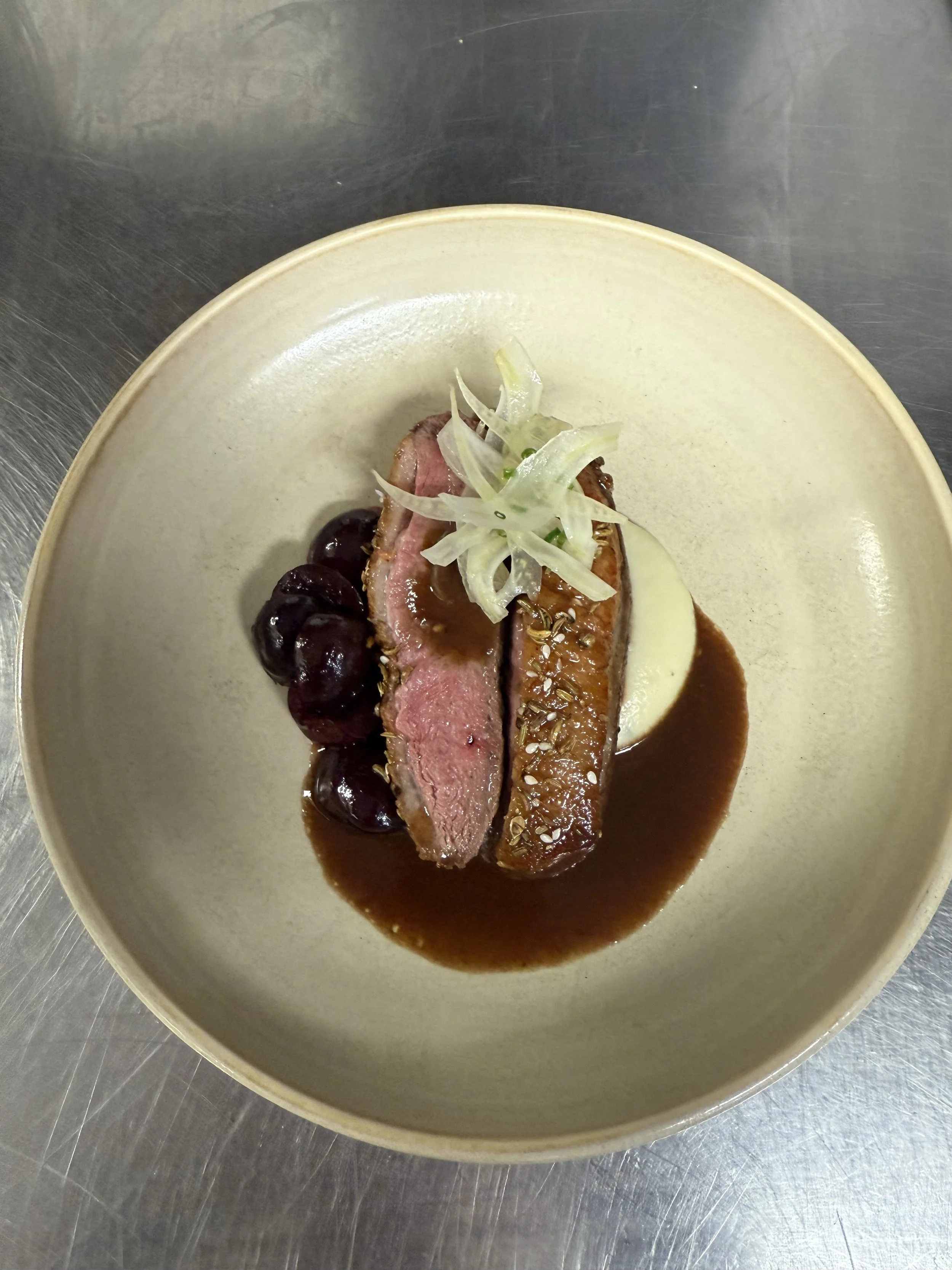 A plate with a piece of cooked meat topped with sliced onions, garnished with dark cherries and covered with a brown sauce.