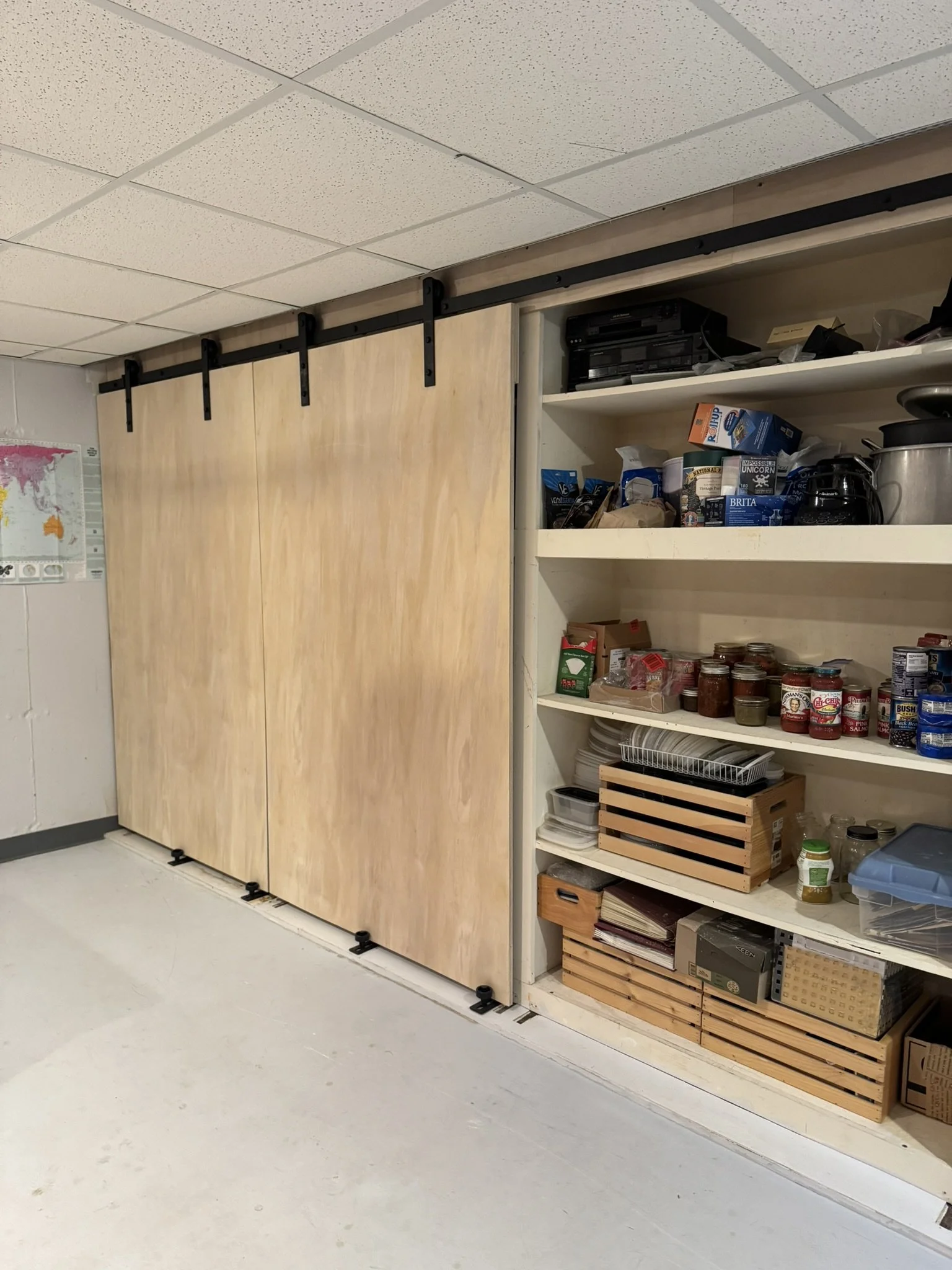 A wooden sliding door on a black metal track, adjacent to a white open shelving unit filled with canned goods, boxes, and kitchen appliances in a room with light-colored flooring and a speckled ceiling.