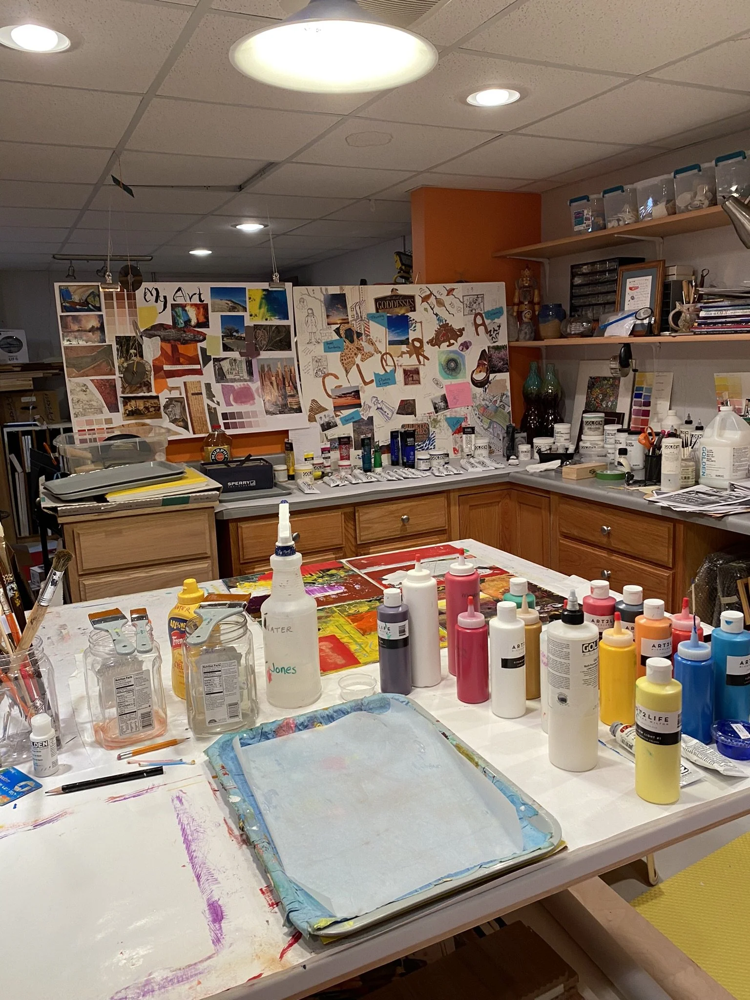 An art studio workspace with paint bottles, brushes, and a large watercolor paper on a table. The background includes a wall with artwork, supplies, and shelves.