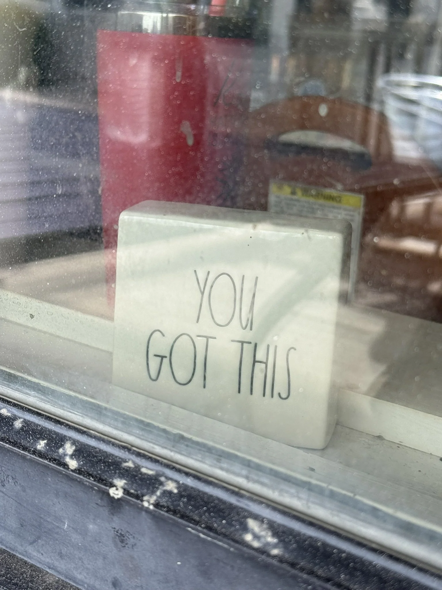 A blurry image taken through a window shows a red cup, part of a sign with the text "YOU GOT THIS" visible, and some other objects.