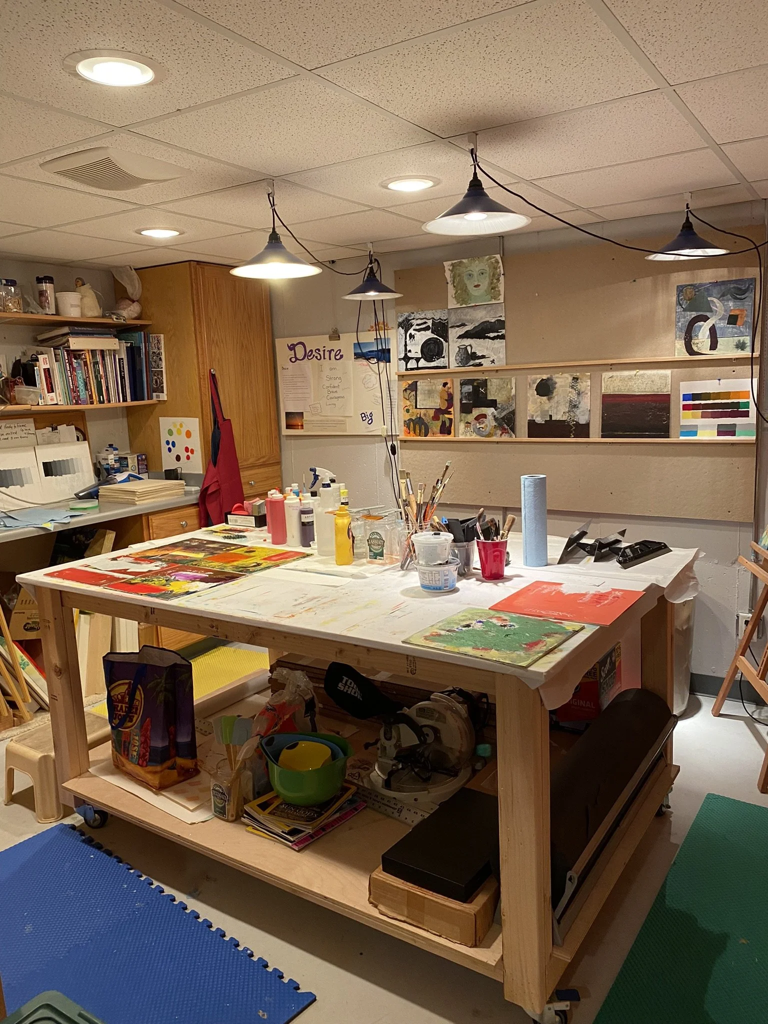 A craft or art studio workspace with a wooden table cluttered with paints, brushes, and artwork. Shelves in the background hold books and supplies, with multiple paintings displayed on the wall and ceiling lights hanging above.