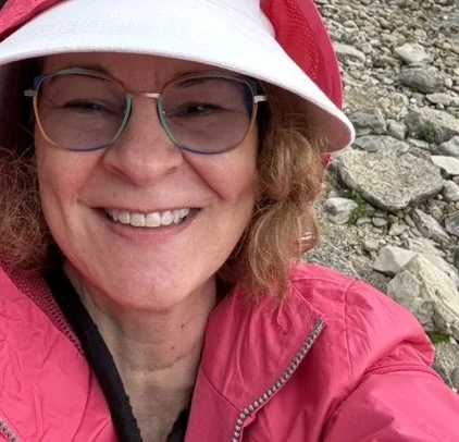 Woman smiling outdoors, wearing glasses, a pink jacket, and a white and red hat, with rocky terrain in the background.