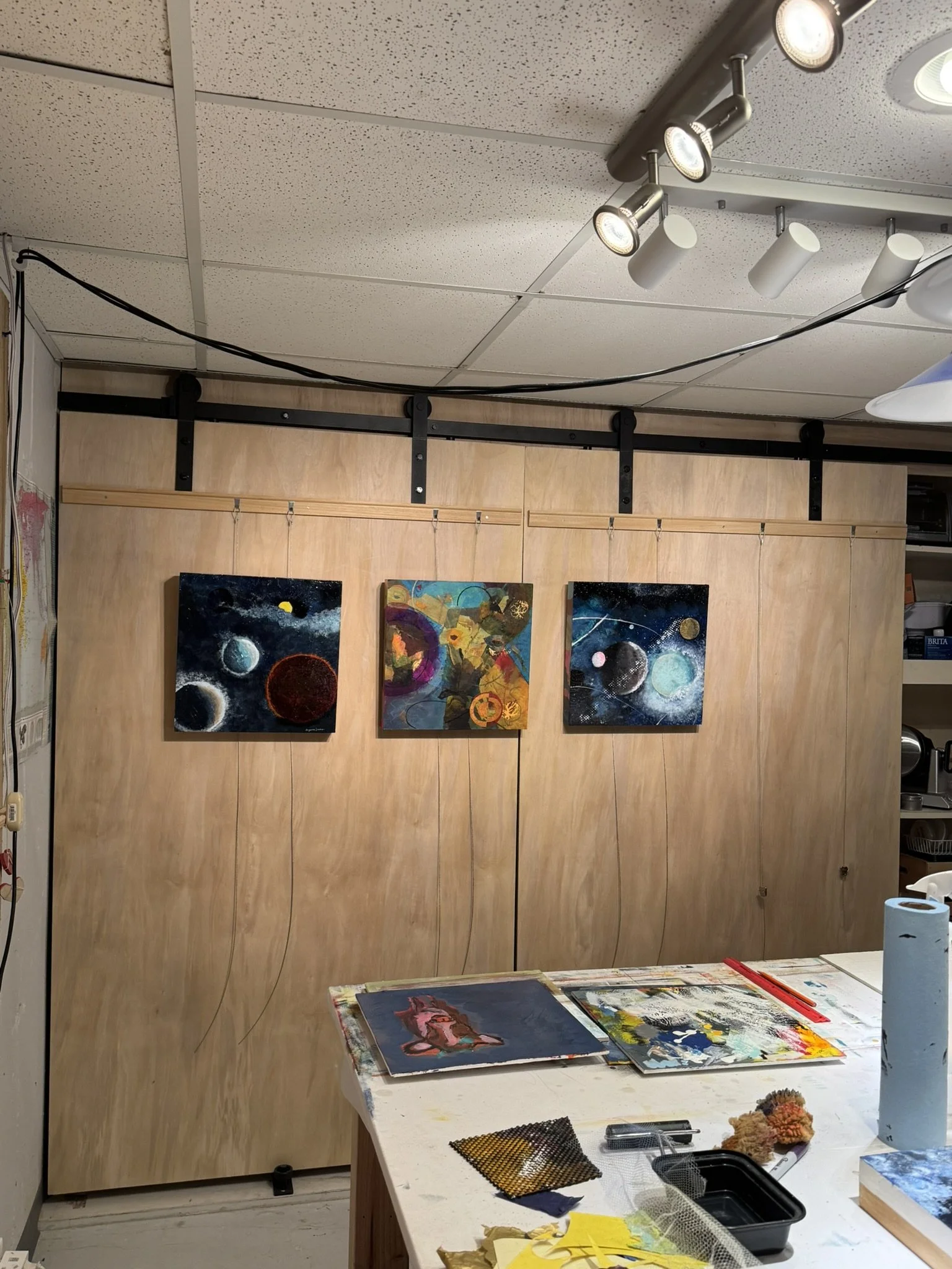 An art studio with paintings of planets on a wooden wall, a worktable with various art supplies including paintings, brushes, and textiles, and ceiling lights illuminating the workspace.