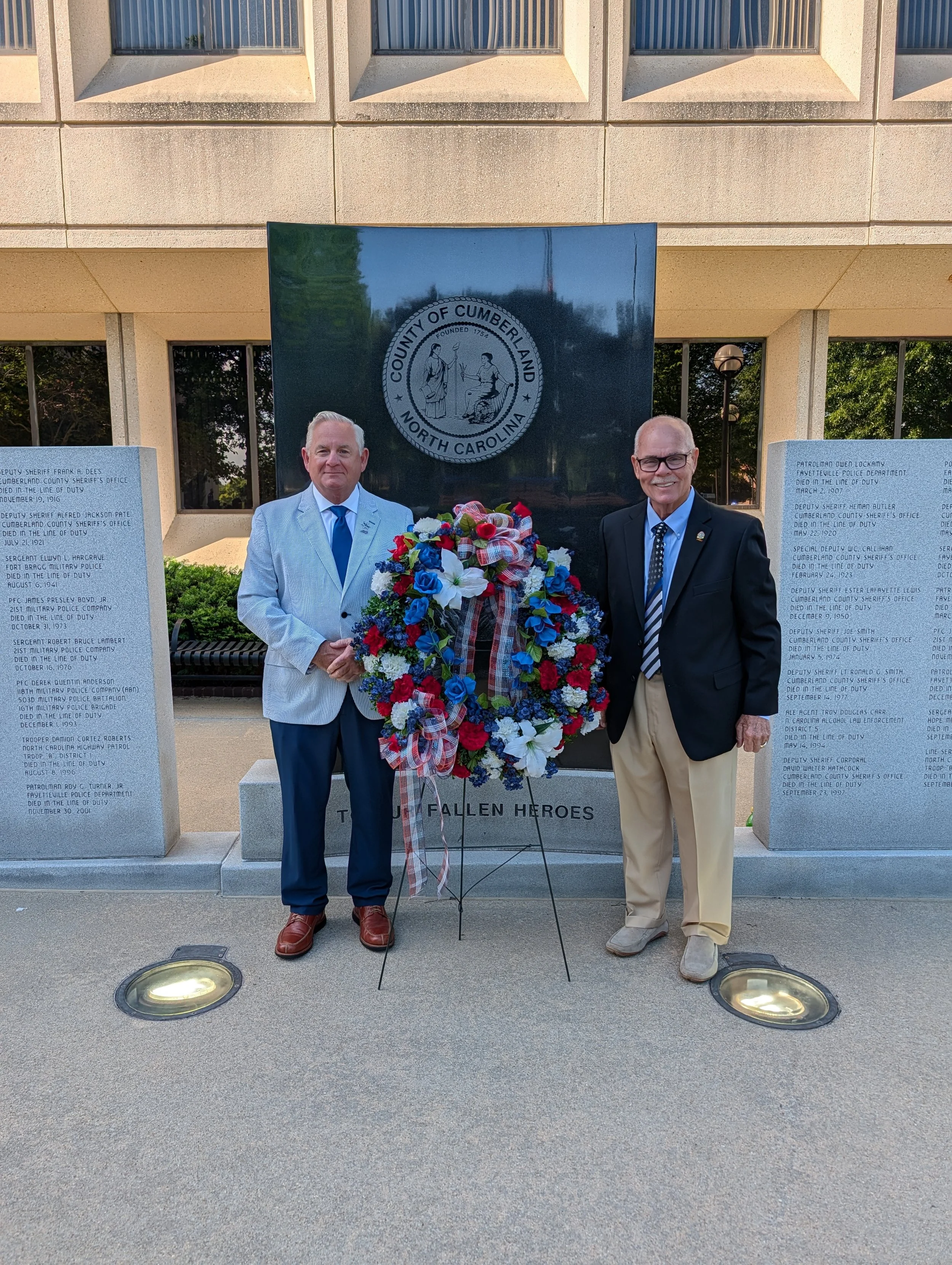 Two elderly men standing outside in front of a memorial wall. One man in a light blue striped suit holds a multicolored wreath of flowers, and the other man in a dark suit and glasses stands beside him, smiling. The memorial wall features plaques with names and dates of fallen heroes from Cumberland County, North Carolina.