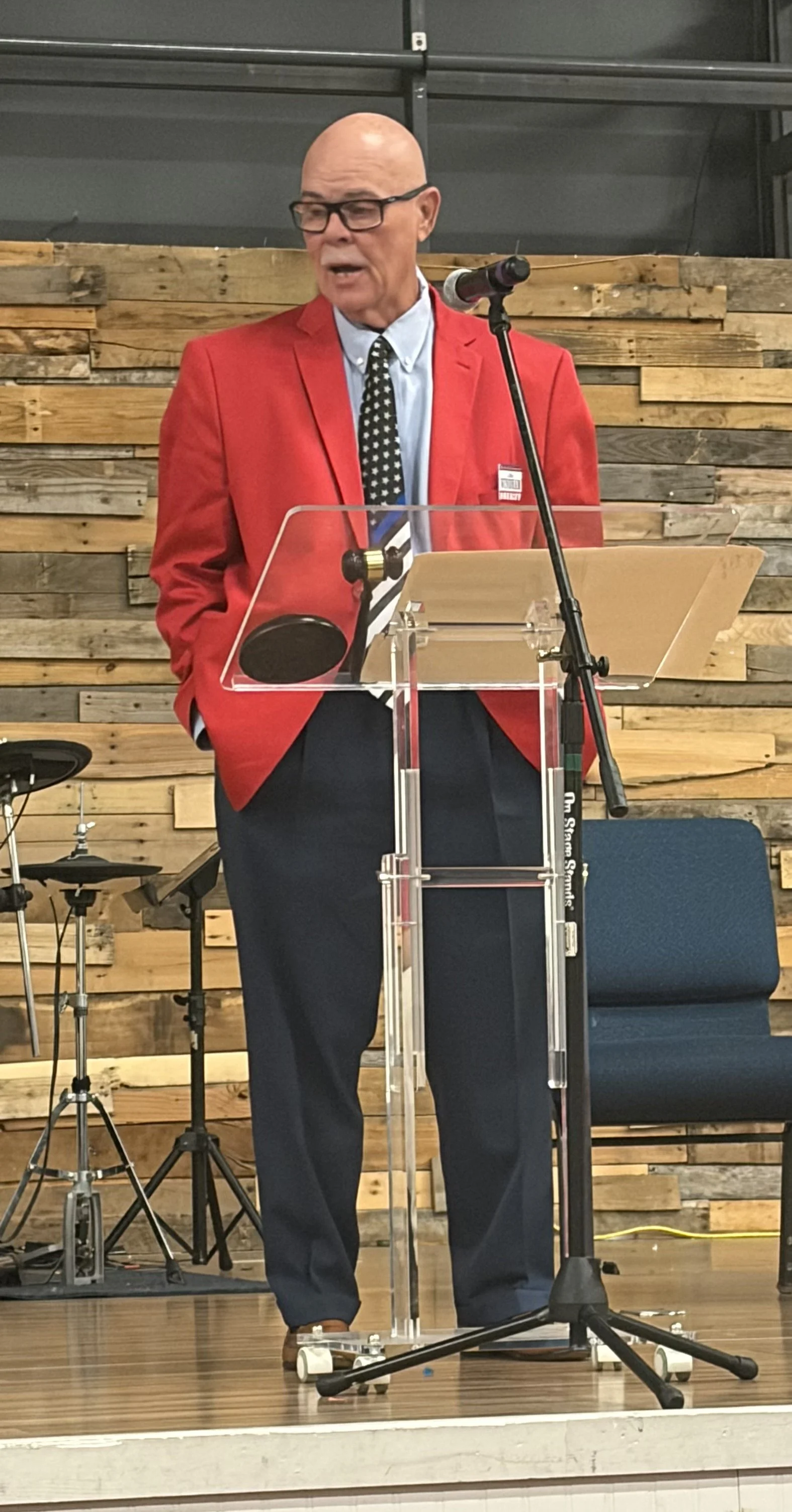 A man wearing a red blazer, glasses, and a black and white polka-dotted tie, speaking at a podium with a microphone, in front of a wooden wall background.