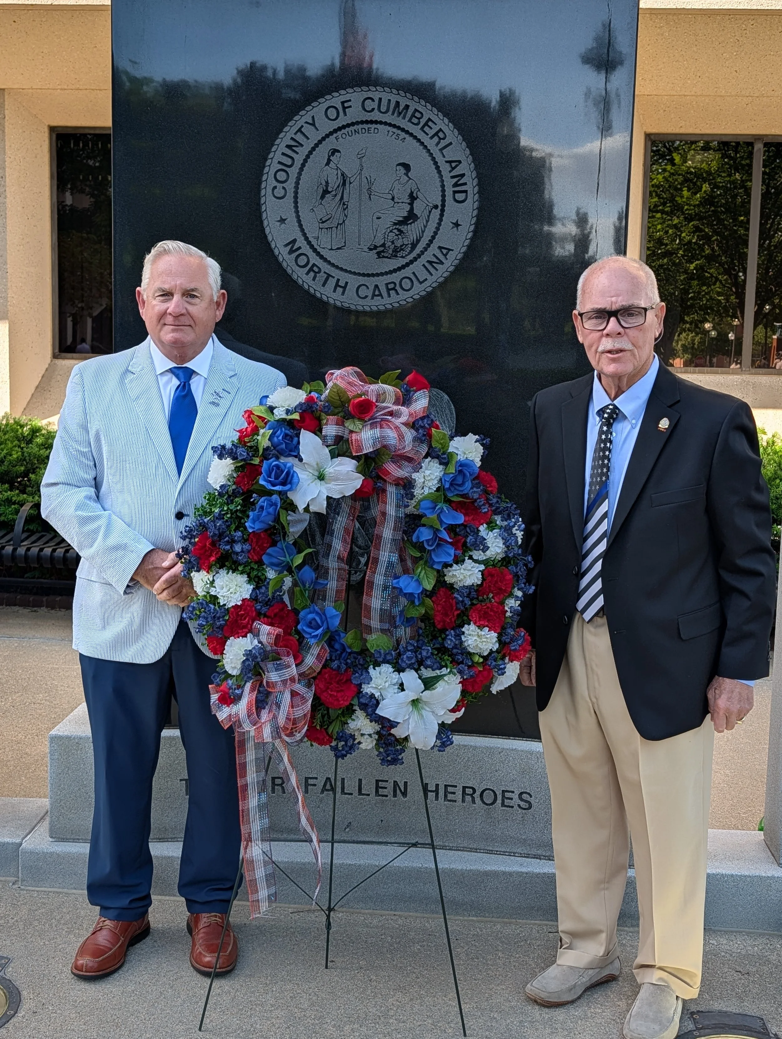 Two men standing beside a memorial with a wreath of flowers. The memorial has a seal with the text 'County of Cumberland North Carolina' and an inscription at the bottom that reads 'For Fallen Heroes'. One man wears a light-colored suit with a blue tie, and the other wears a black jacket, light-colored pants, and a patterned tie.
