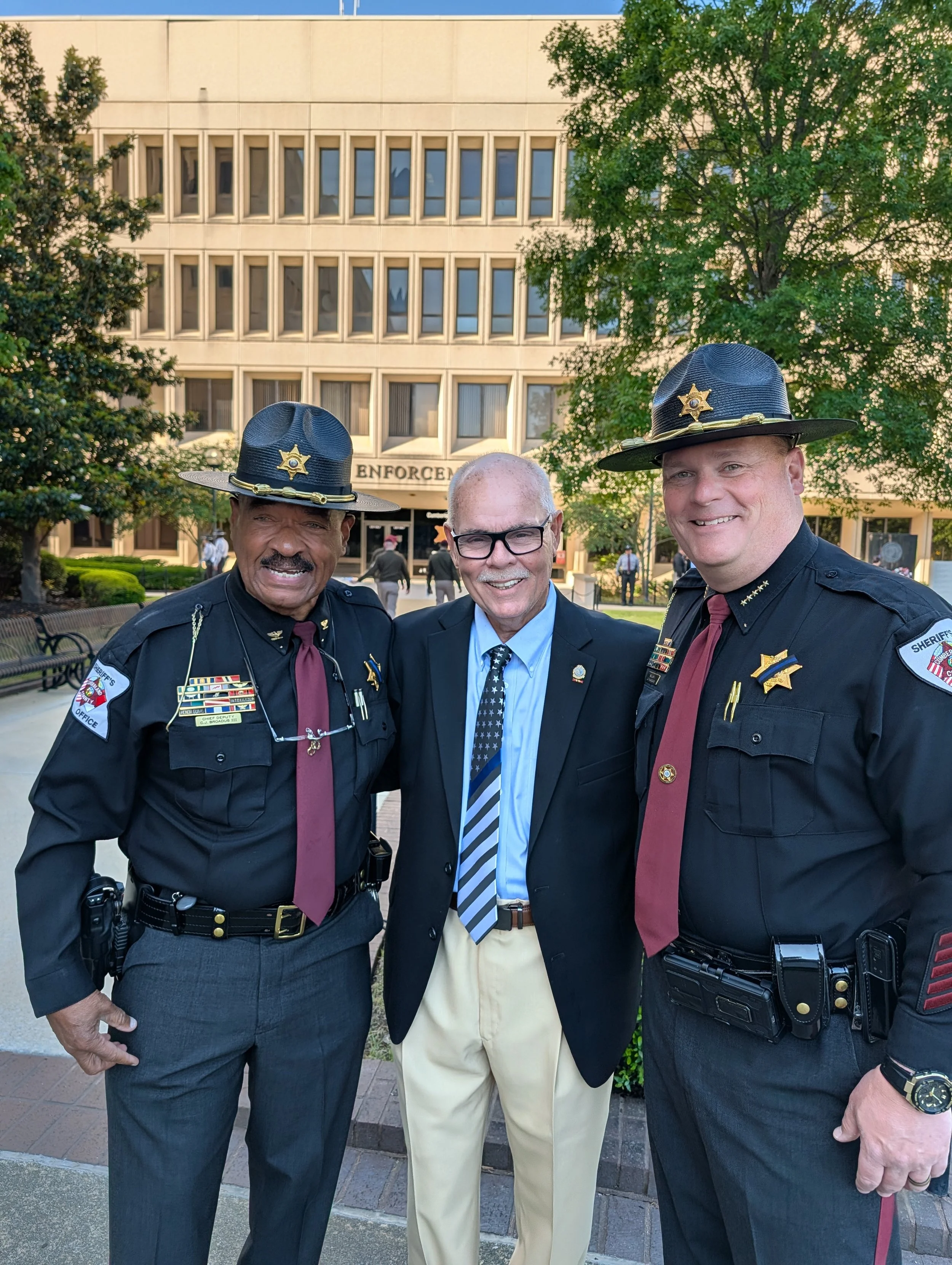Three men standing outdoors in front of a beige building with trees. Two of the men are police officers wearing black uniforms and hats with sheriff badges. The middle man is dressed in a black blazer, blue shirt, and a tie with stars. They are smiling and posing for the photo.
