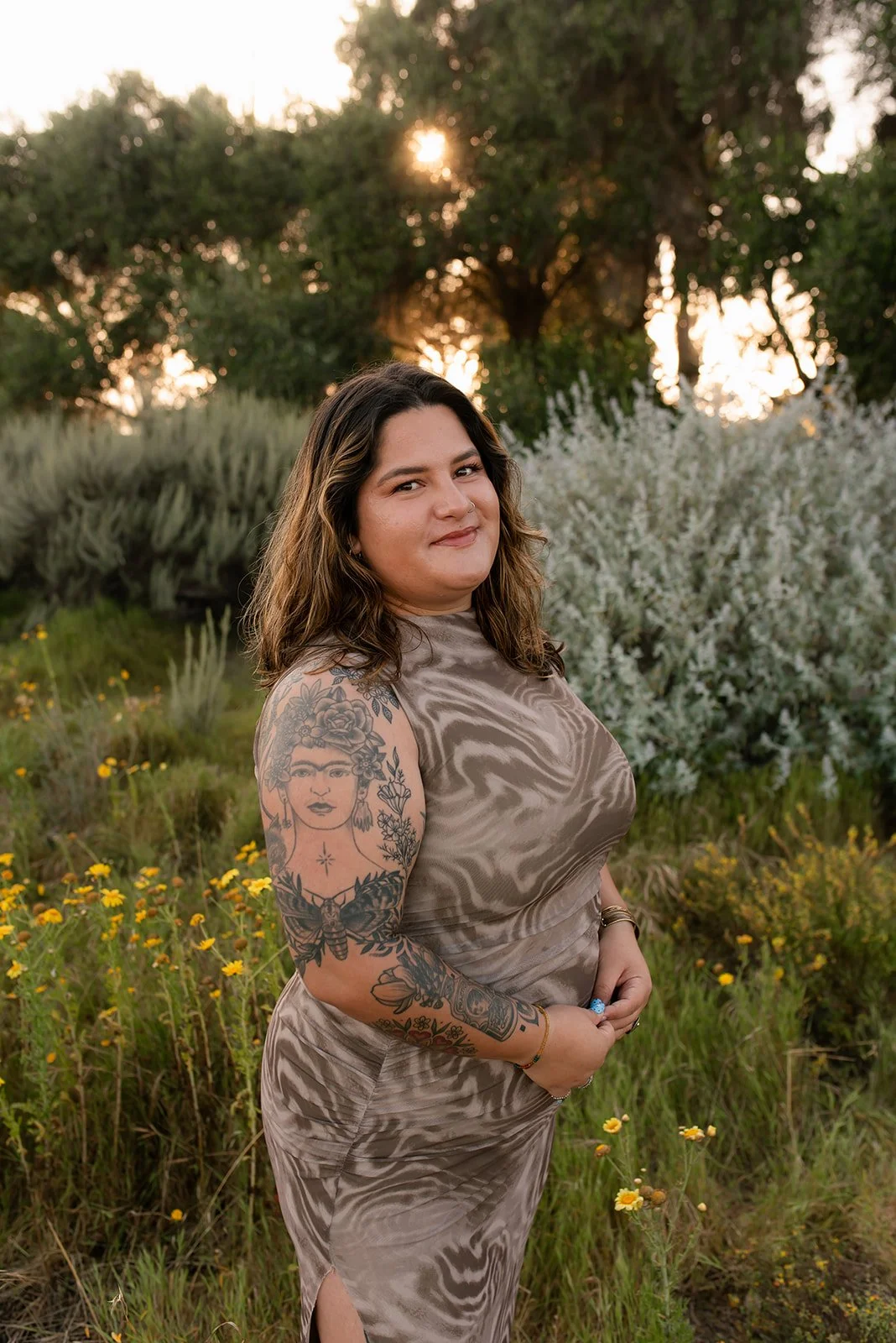 A woman with long dark hair standing outdoors during sunset, wearing a patterned dress, with visible tattoos on her left arm, surrounded by greenery and wildflowers.