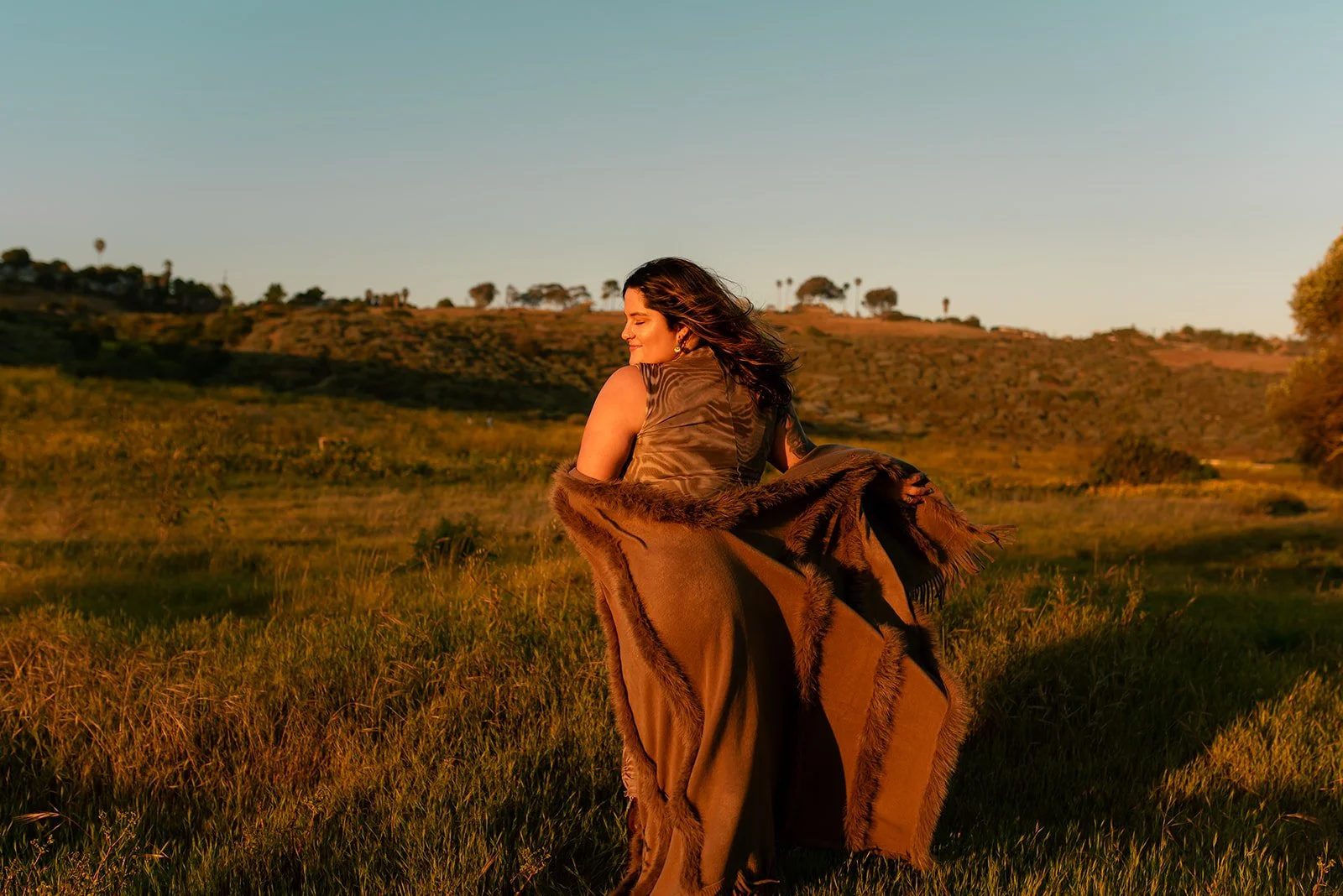 A woman with dark hair wearing a sleeveless top, holding a brown fur-lined coat, standing in a field during sunset or sunrise with rolling hills and a few trees in the background.