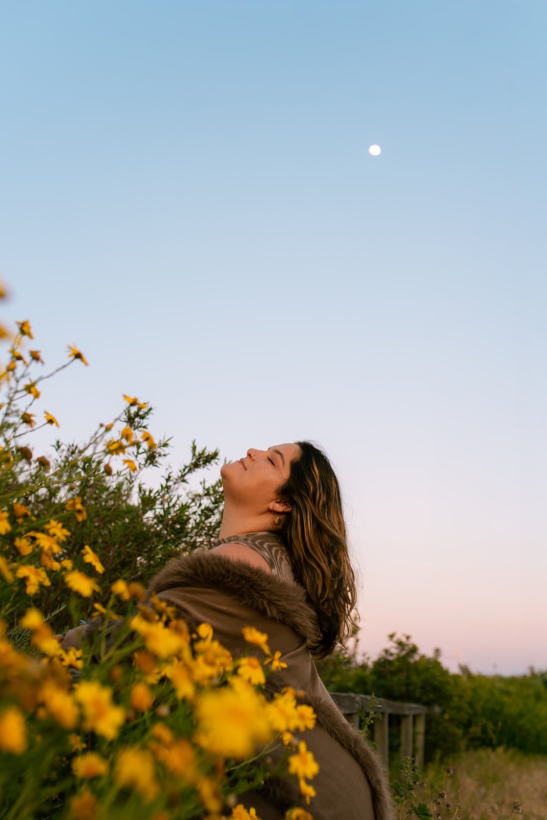 A woman with curly hair standing among yellow flowers outdoors at sunset, with her eyes closed and a peaceful expression, under a clear blue sky with the moon visible.