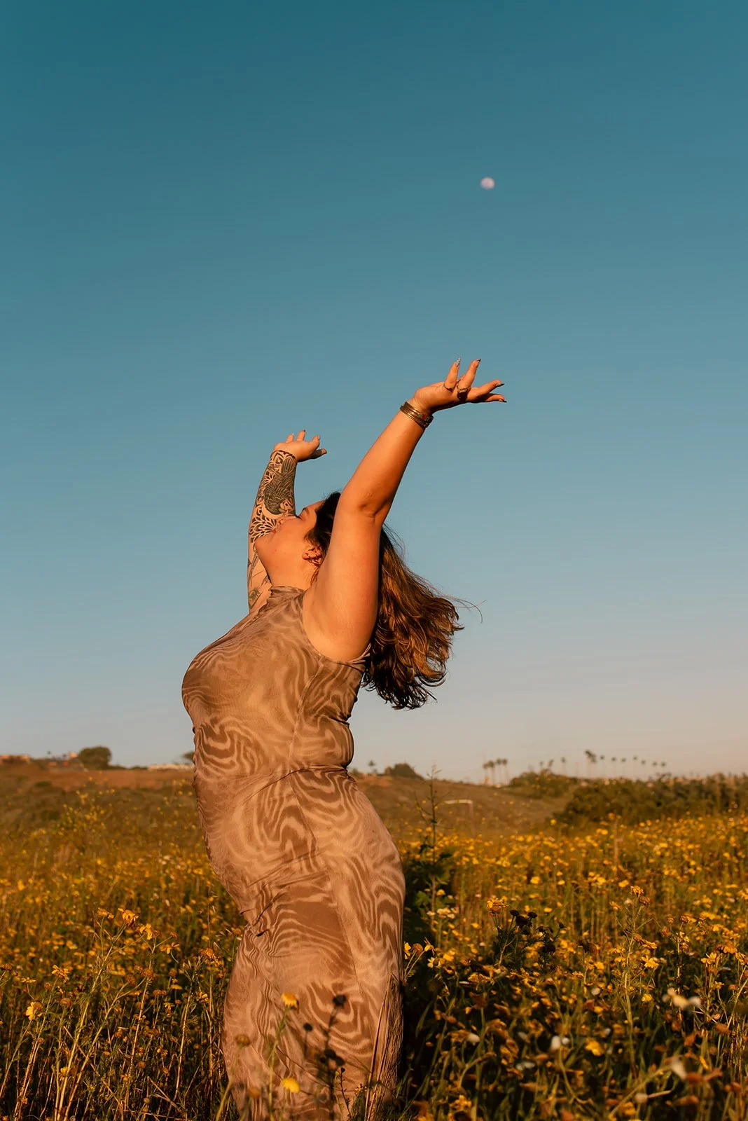 A woman standing in a field of yellow flowers, raising her arms towards the sky with her face turned upward, under a clear blue sky with the moon visible in the distance.