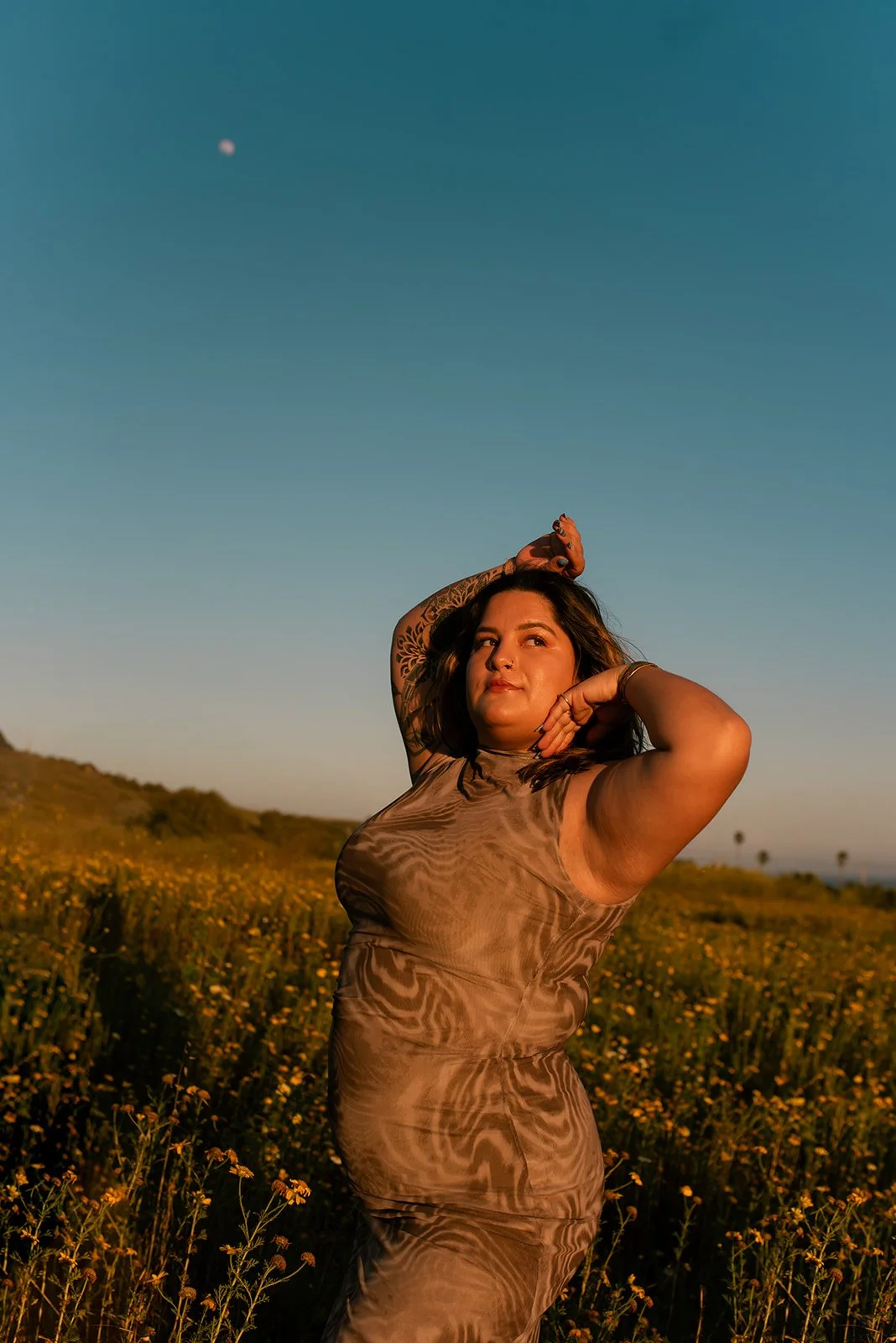 A woman with tattoos on her arms posing in a field of yellow flowers during golden hour, with a blue sky and the moon visible overhead.