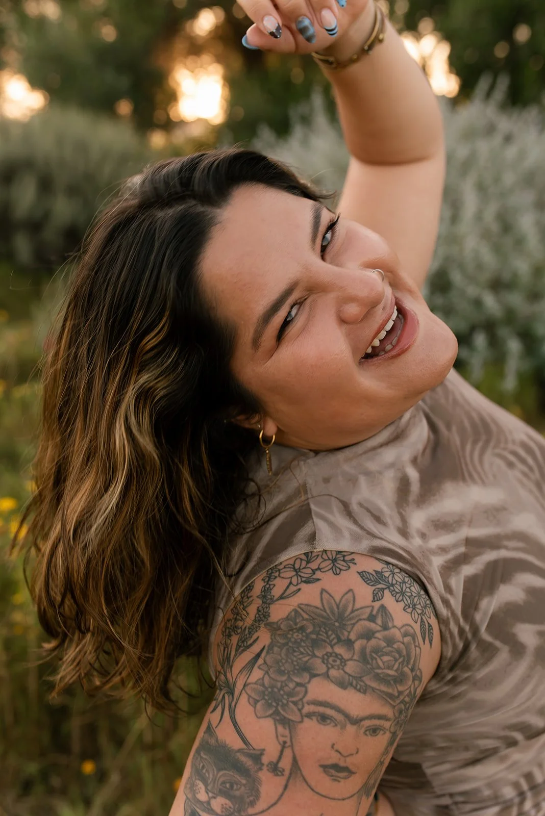 A woman with wavy, brown hair and multiple tattoos, smiling and lying outdoors in a natural setting during sunset.