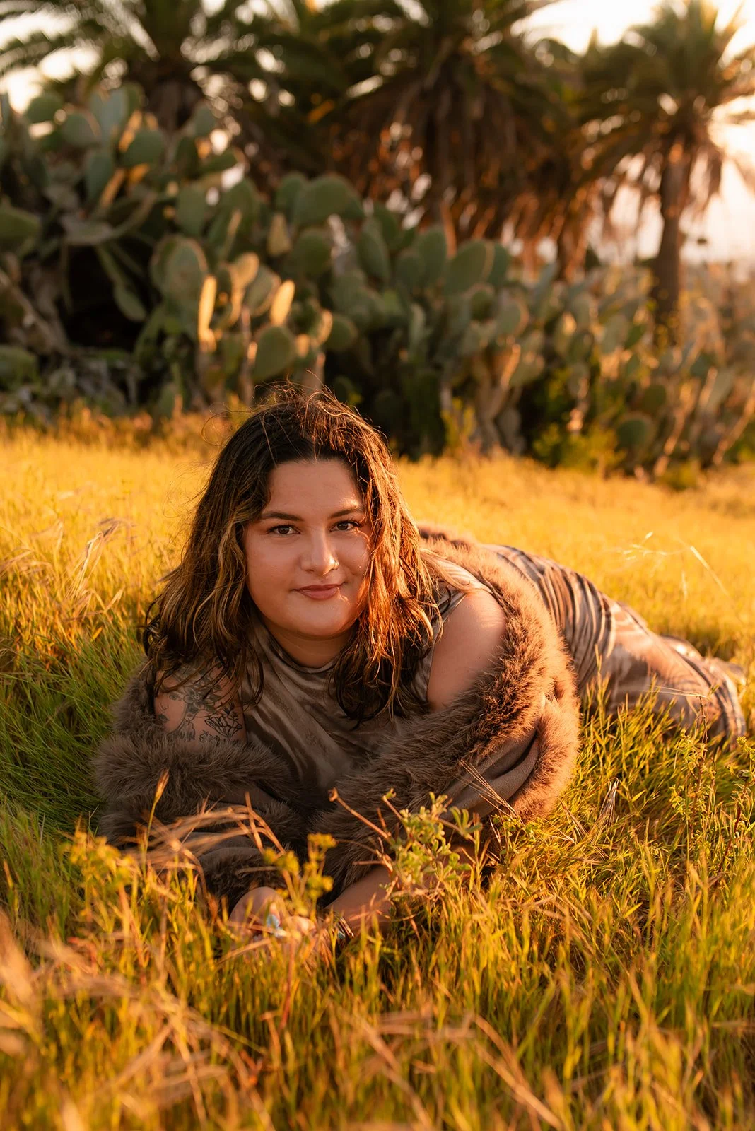 A woman with wavy hair lying on her stomach in a field with tall grass during sunset, wearing a fur coat and camouflage clothing, with large cactus plants in the background.
