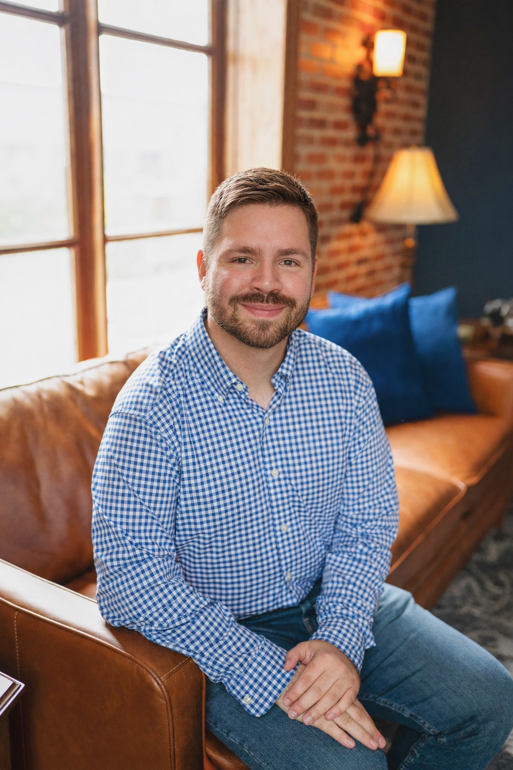 A man with short brown hair, a beard, and a mustache is sitting on a brown leather couch, smiling. He is wearing a blue and white checkered shirt and jeans. The background shows a window, a brick wall, a wall-mounted lamp, and a table lamp with a beige shade, with blue cushions on the couch.