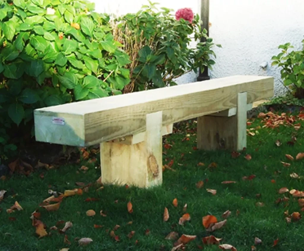 Wooden bench in front of green leafy bushes with pink flowers, on a grassy yard scattered with fallen autumn leaves.