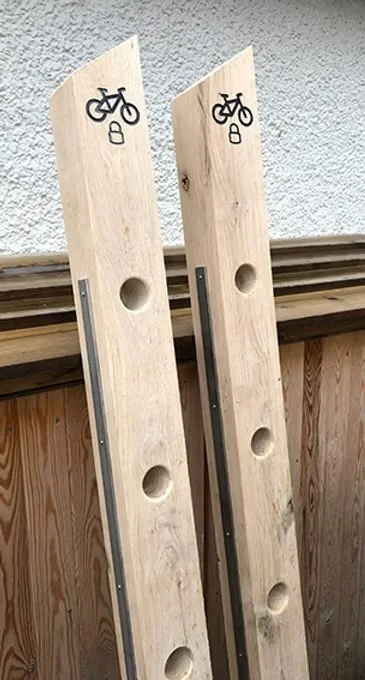 Two wooden skateboard decks with bike and lock symbols on top, and circular holes and metal rails on the sides, leaning against a wall.