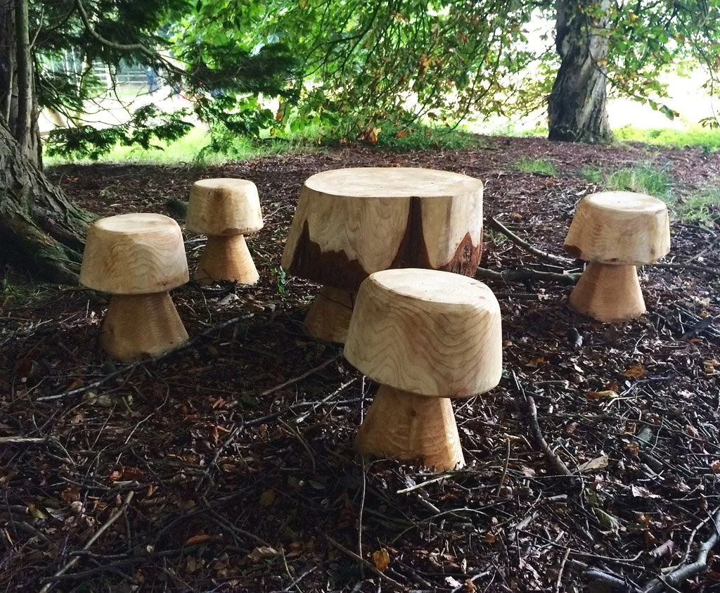 Wooden mushroom-shaped stools and a table arranged outdoors on forest ground with trees and green foliage in the background.