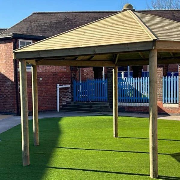 Wooden gazebo with a pointed roof in a grassy area, with a red brick building and a blue fence in the background.
