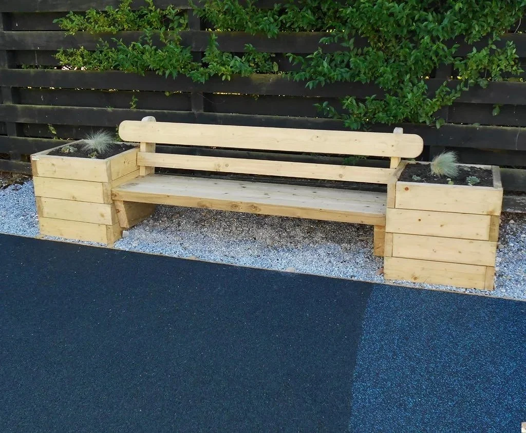 Wooden outdoor bench with side planters filled with soil and small plants, set against a black wooden fence and green shrub background, on a gravel and asphalt surface.