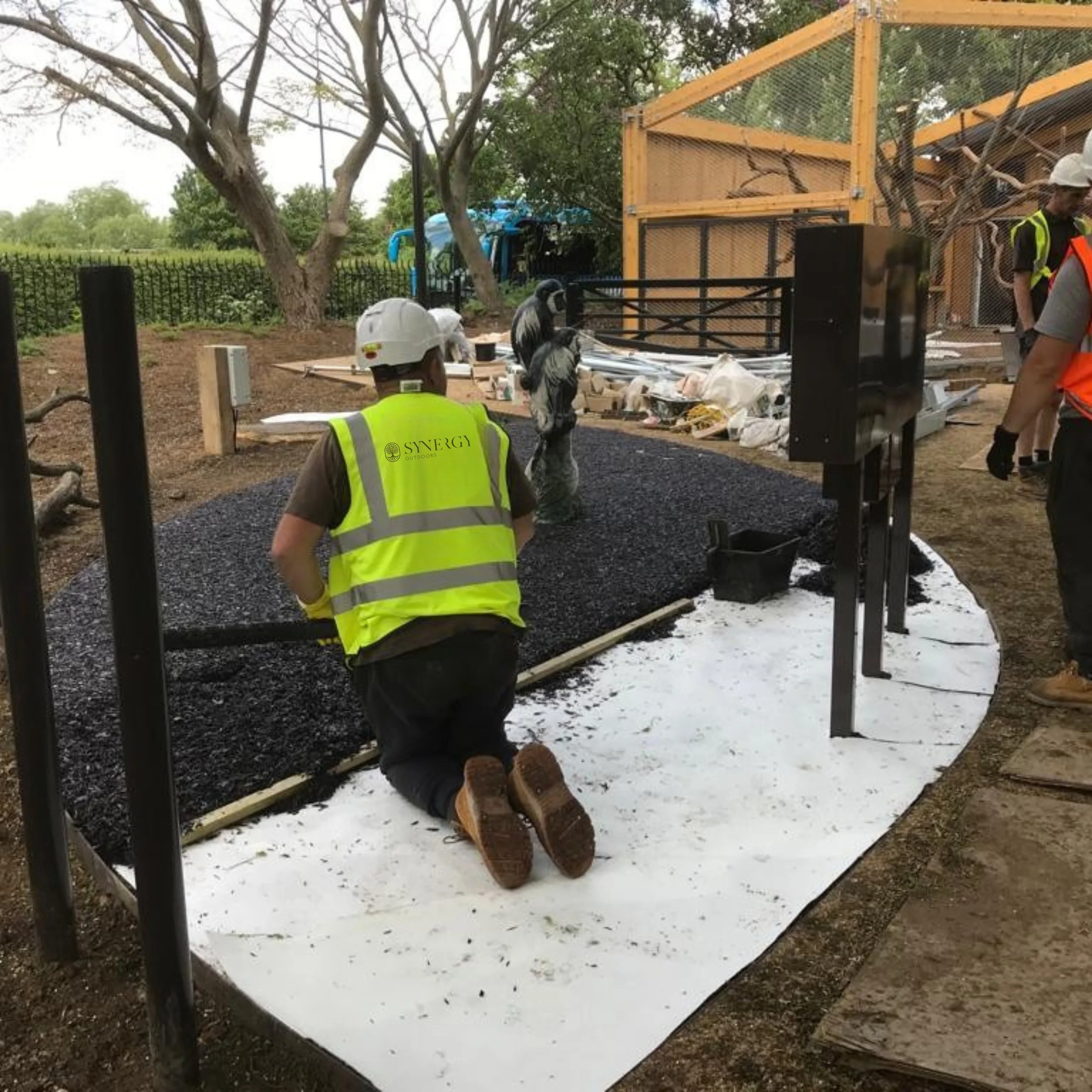 Construction workers installing a black gravel pathway, with one kneeling in the gravel and others standing nearby, surrounded by construction materials and machinery outdoors.