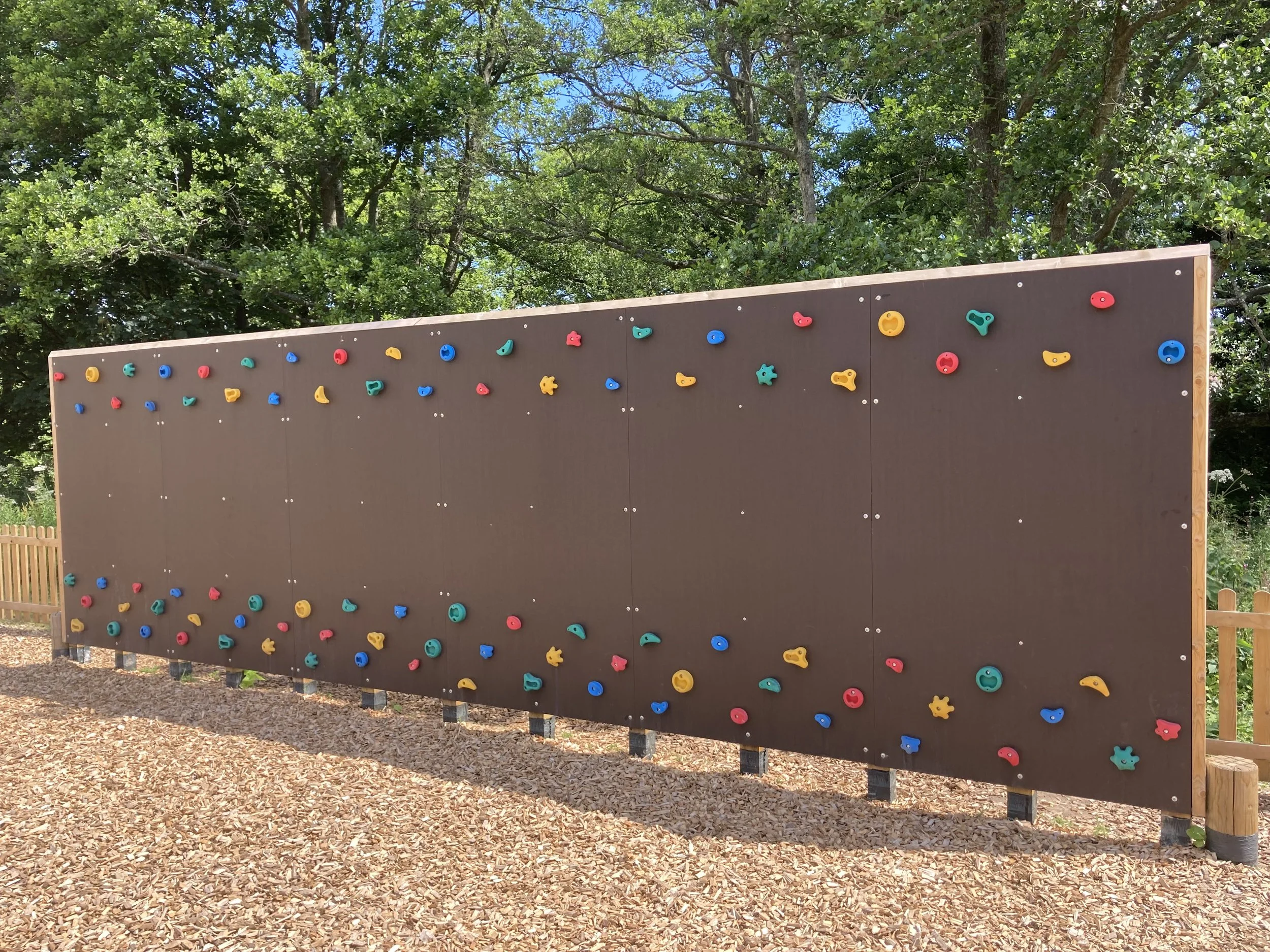 Outdoor climbing wall with colorful holds, set against green trees, on a bed of wood chips.
