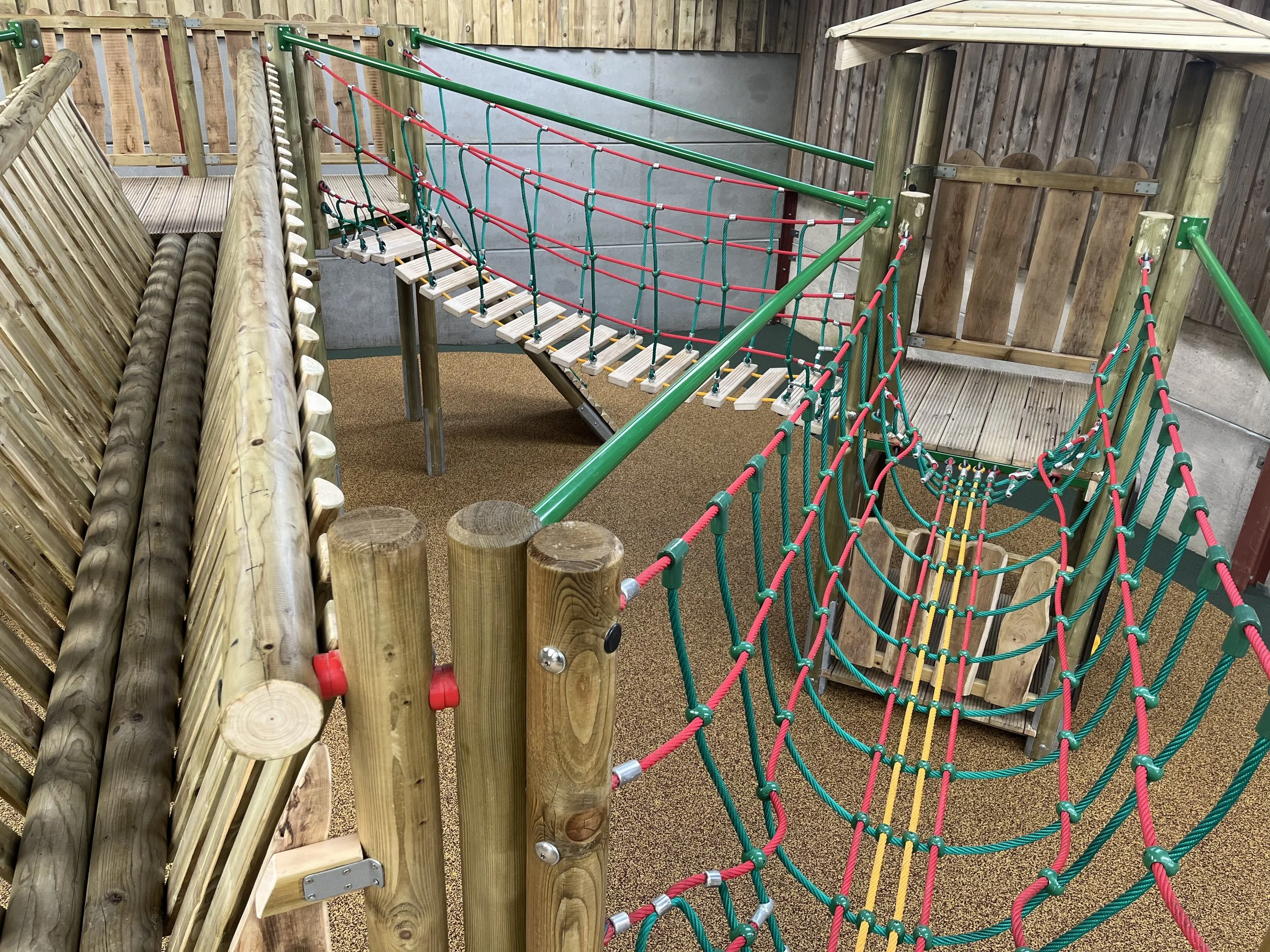 Indoor wooden playground with rope bridges and tunnels, surrounded by wooden fencing and walls.