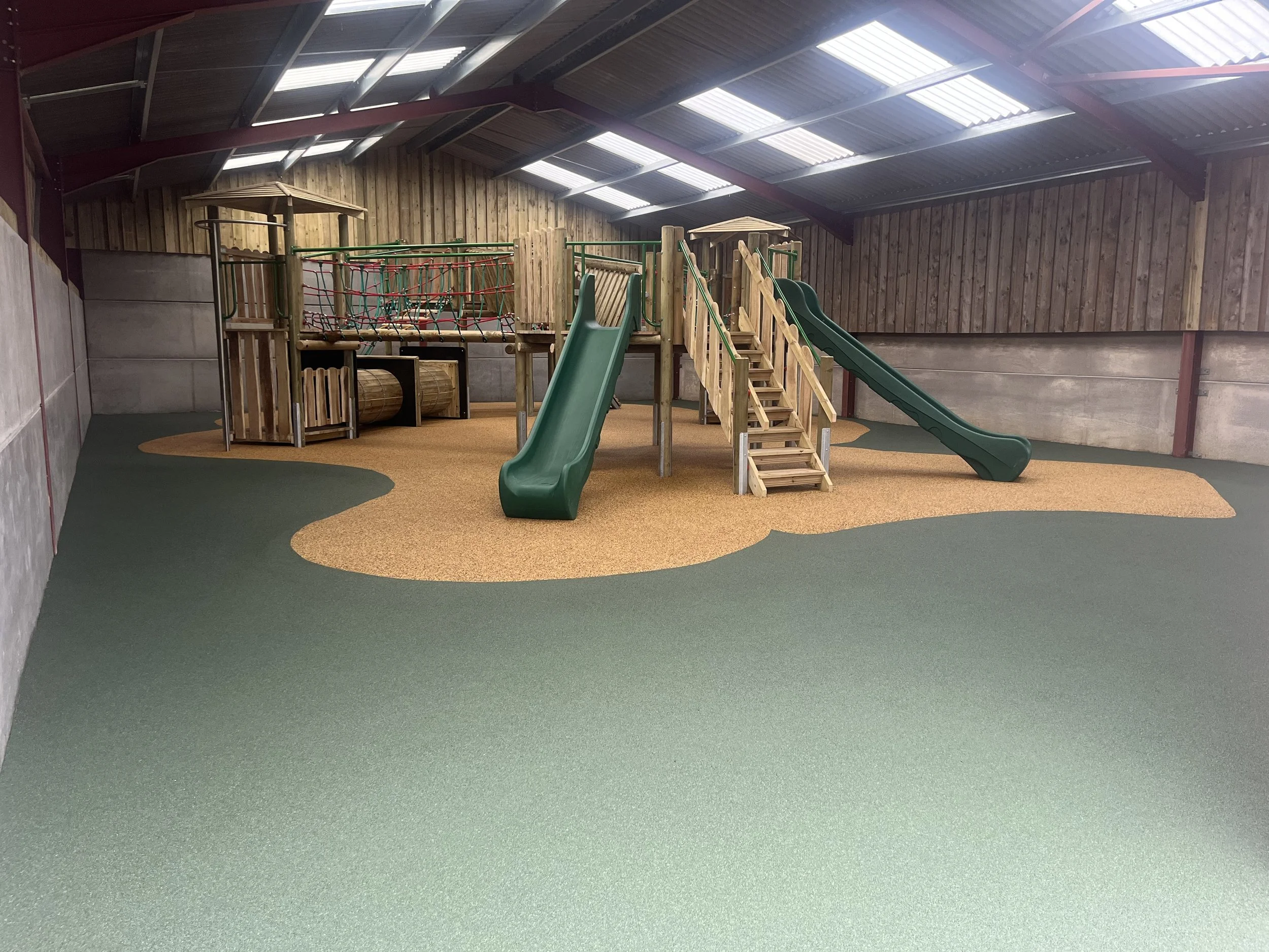Indoor children's playground with wooden play structure, green slides, and climbing net, on a rubberized floor with green and yellow sections.