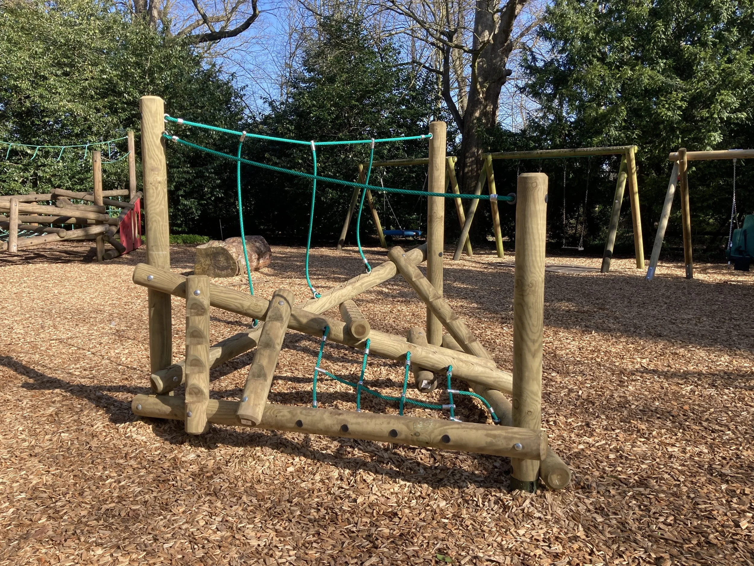 Wooden playground structure with climbing elements and ropes, surrounded by wood chips, in a park with trees in the background.