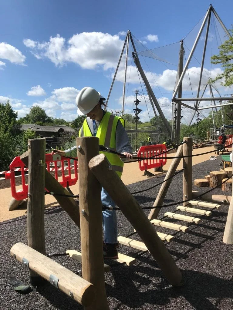 A person in a white safety helmet and yellow safety vest walking on a wooden bridge in a playground or park area, with a blue sky and clouds overhead.