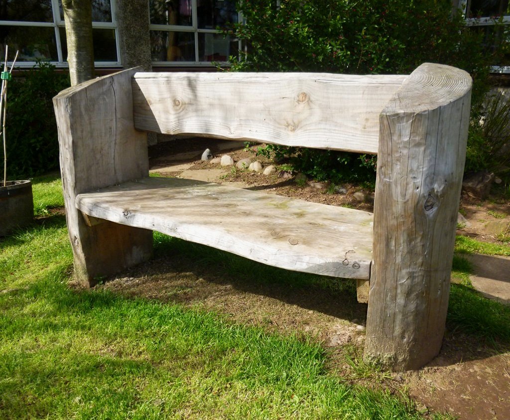An outdoor wooden bench made from rough, natural logs, situated on a grassy area with a building and bushes in the background.