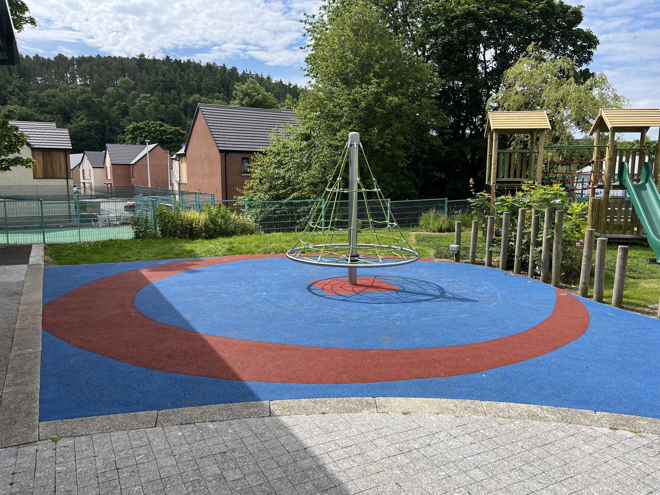 Playground with a colorful rubber safety surface, a metal spinning toy in the center, and wooden play structures with slides in the background, surrounded by a fence and greenery.