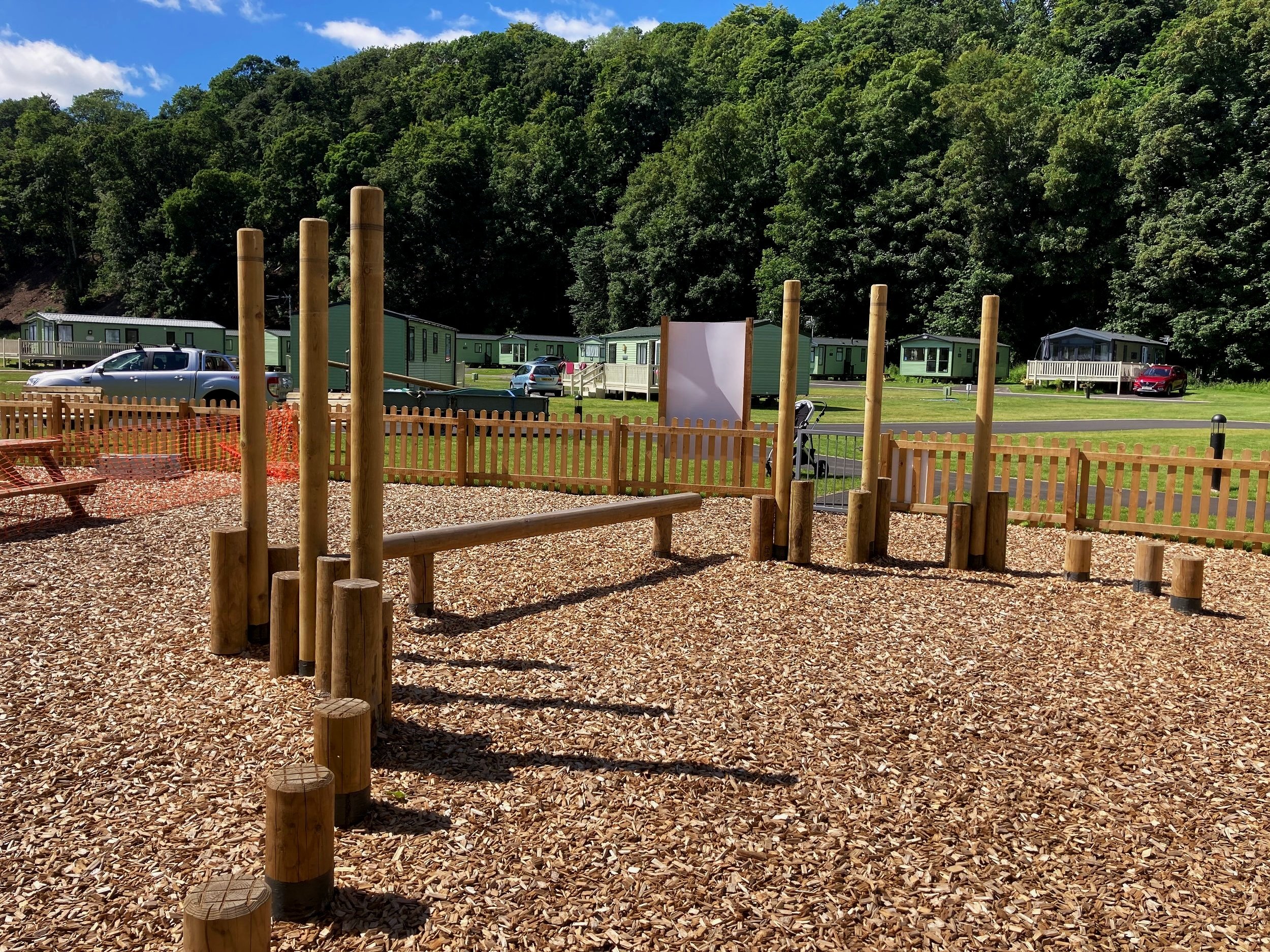 A playground with wood chips, a wooden fence, and vertical wooden posts, set against a background of parked cars, green mobile homes, and a wooded hillside.