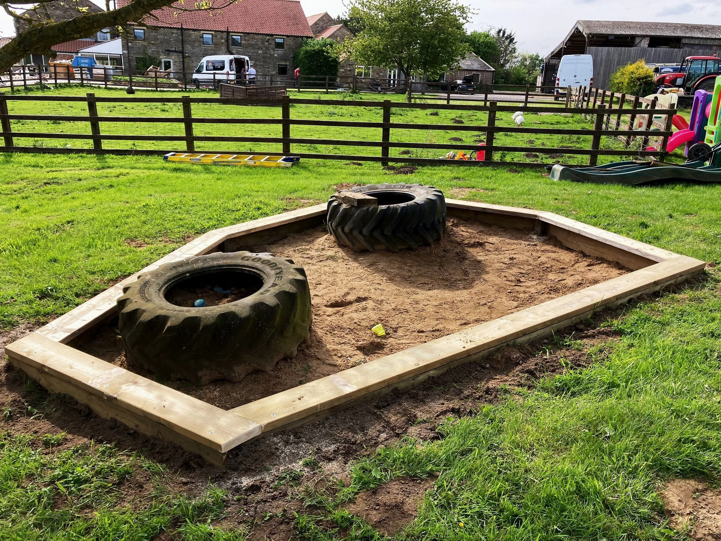 A sandbox with two large tractor tires inside, set on a grassy field surrounded by a wooden frame. In the background, there are fences, buildings, and playground equipment.