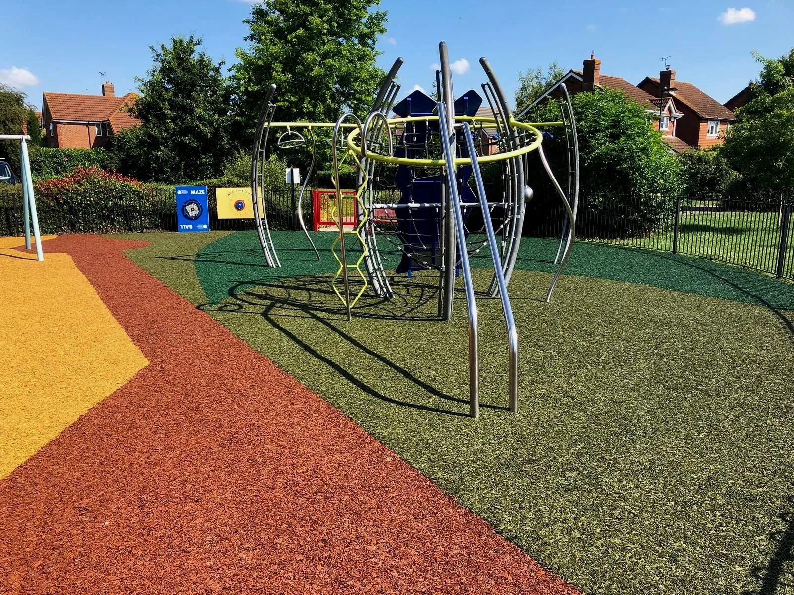 Colorful playground with metal climbing structure, maze panels, and a paved walk path, surrounded by greenery and houses in the background.