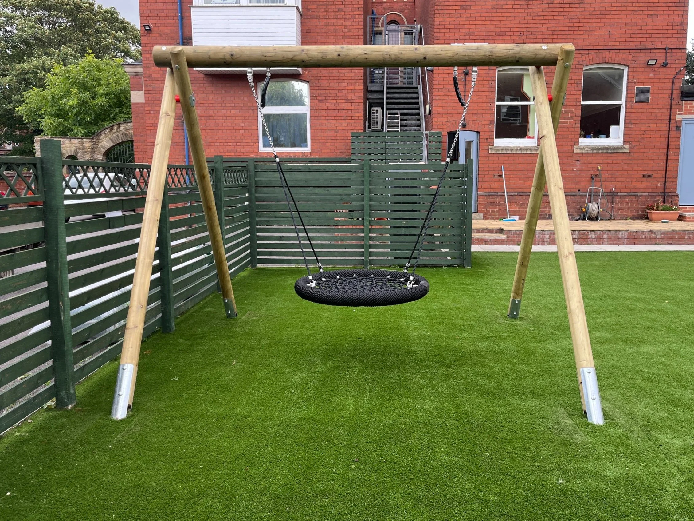 A backyard with a wooden swing set, featuring a circular black net swing, on a green artificial grass surface enclosed by a green privacy fence, with a brick building and windows in the background.