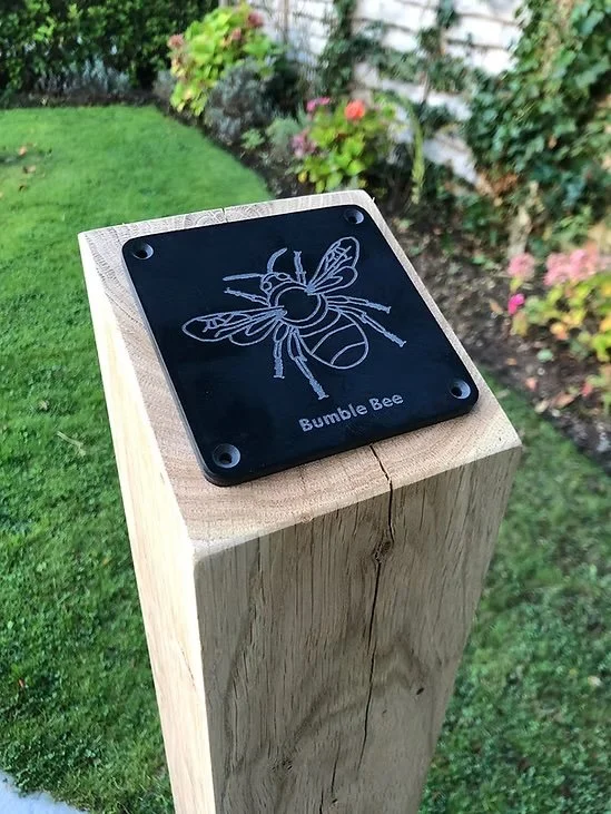 A wooden post with a black metal plaque engraved with a bee illustration and the words "Bumble Bee" outside in a garden with green grass and flowering plants in the background.