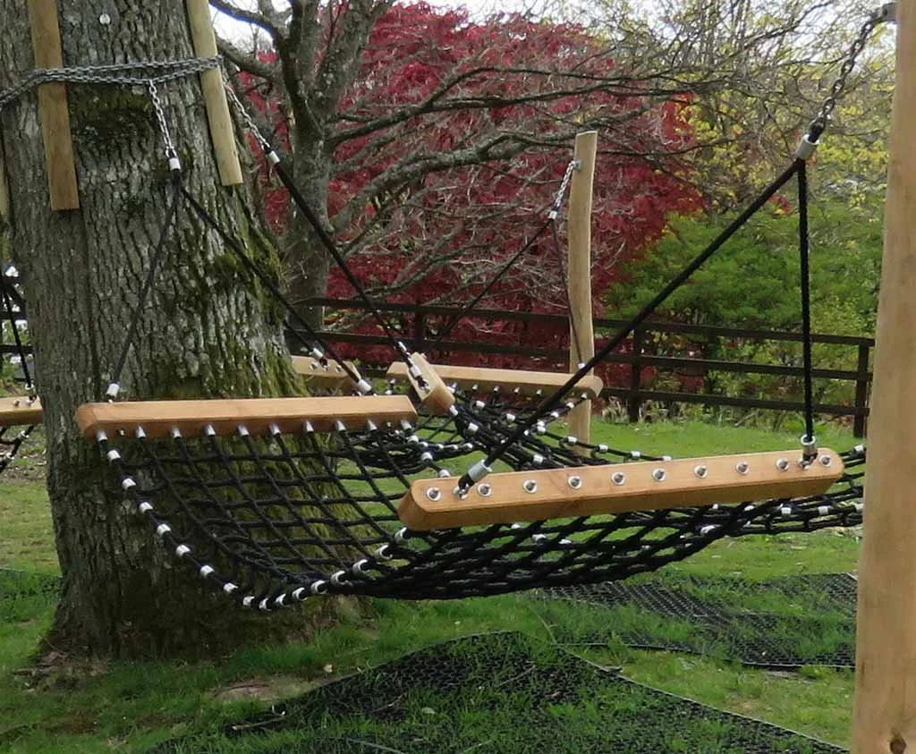 A hanging tree swing with a netted seat, wooden support beams, and ropes, attached to a large tree in a grassy area with trees in fall colors in the background.