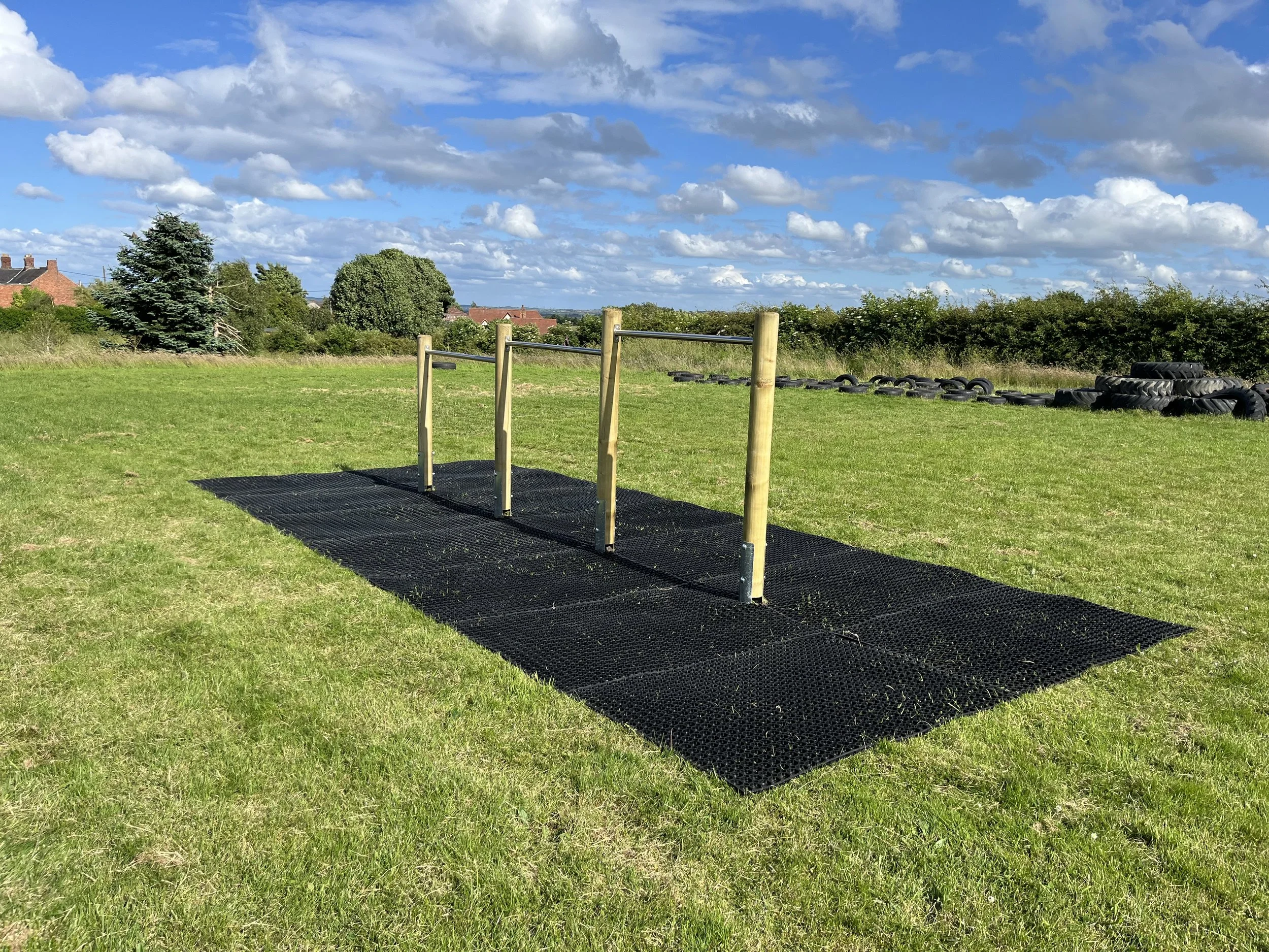 Outdoor fitness obstacle course with wooden bars and black rubber mat, in a grassy field under a partly cloudy sky.